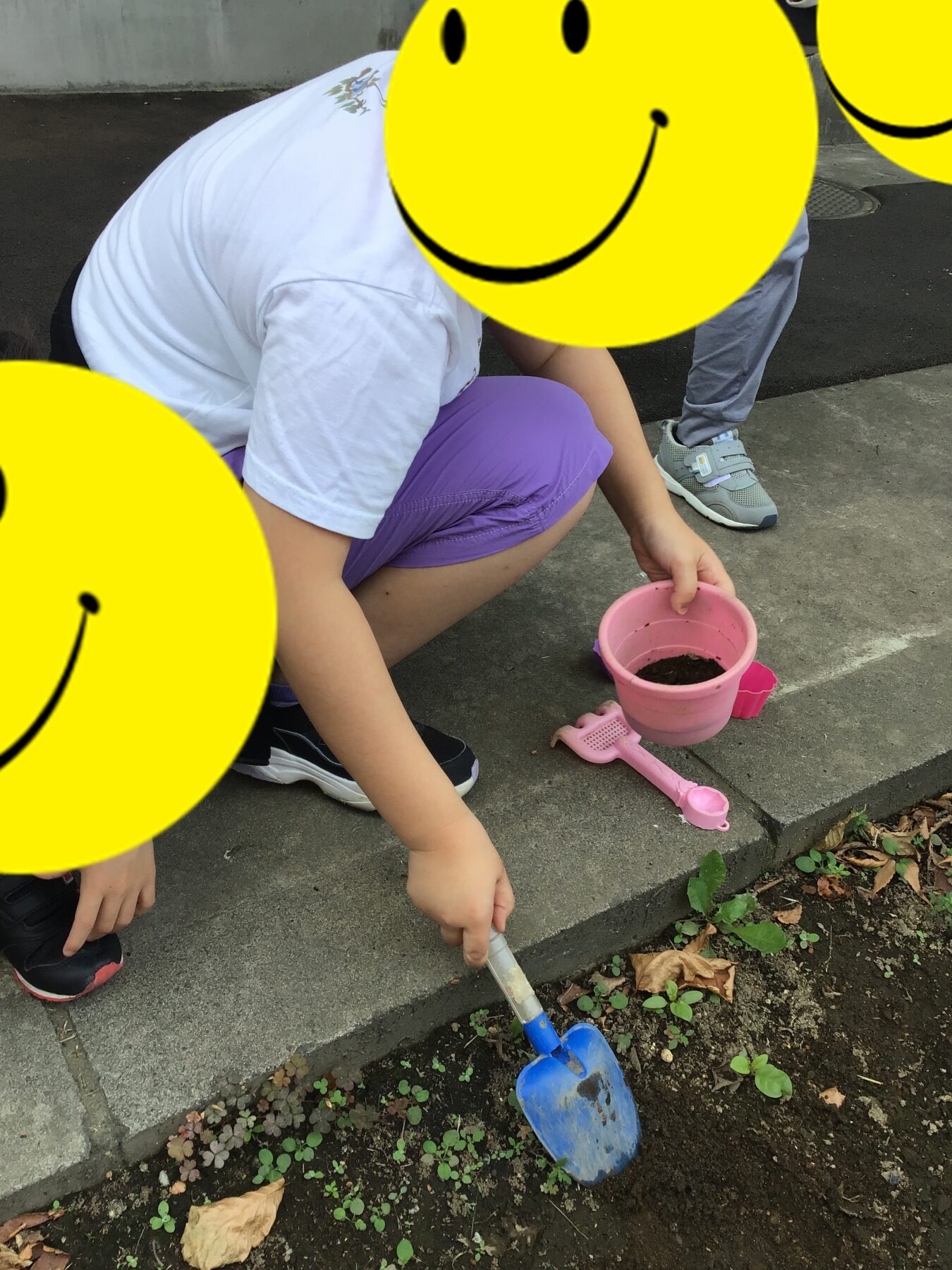 Children kneeling on a curb, digging soil with small gardening tools and a pink bucket nearby.