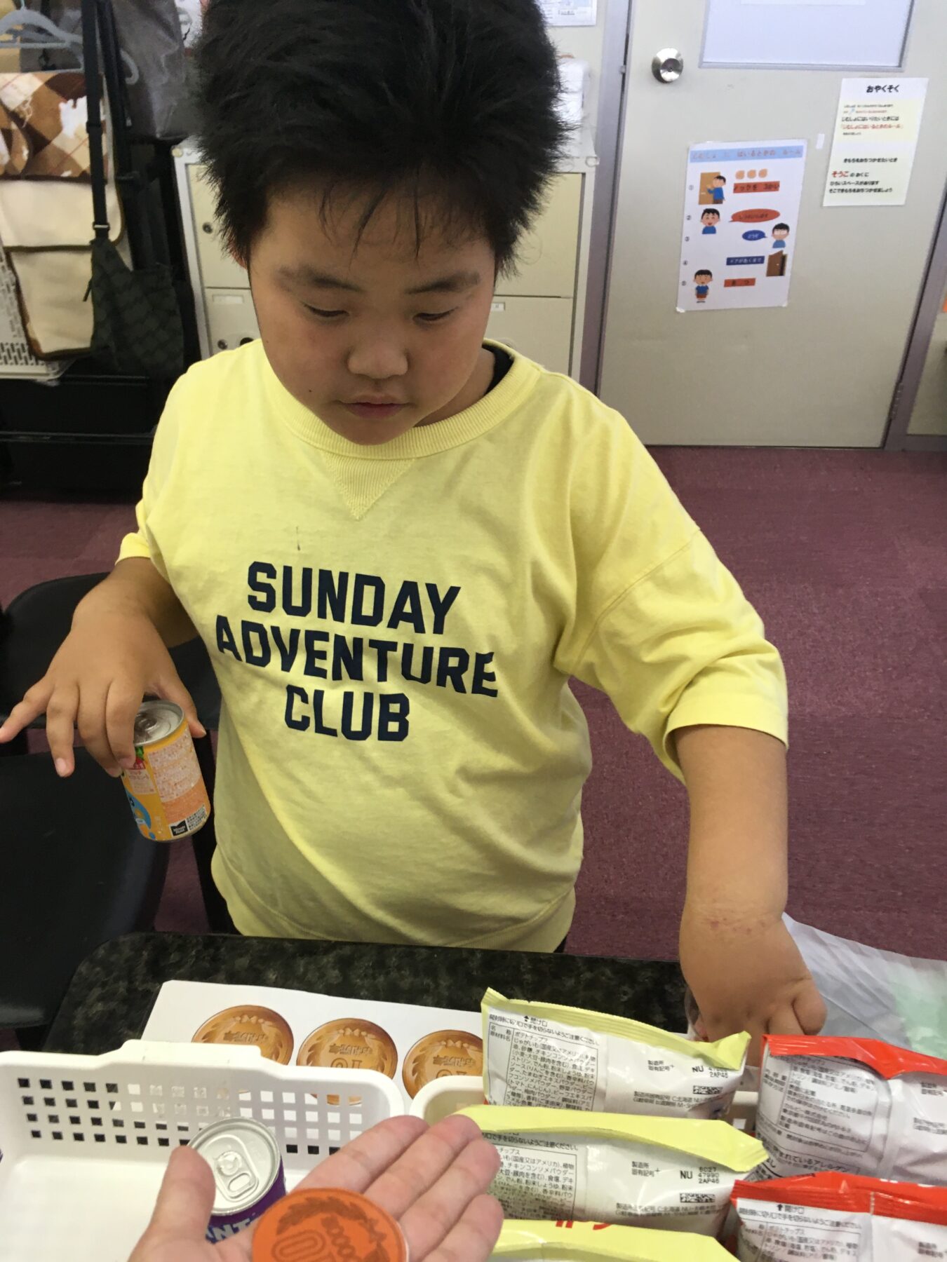 Young boy in a yellow shirt (Sunday Adventure Club) standing at a counter with snacks, looking down at his hands.