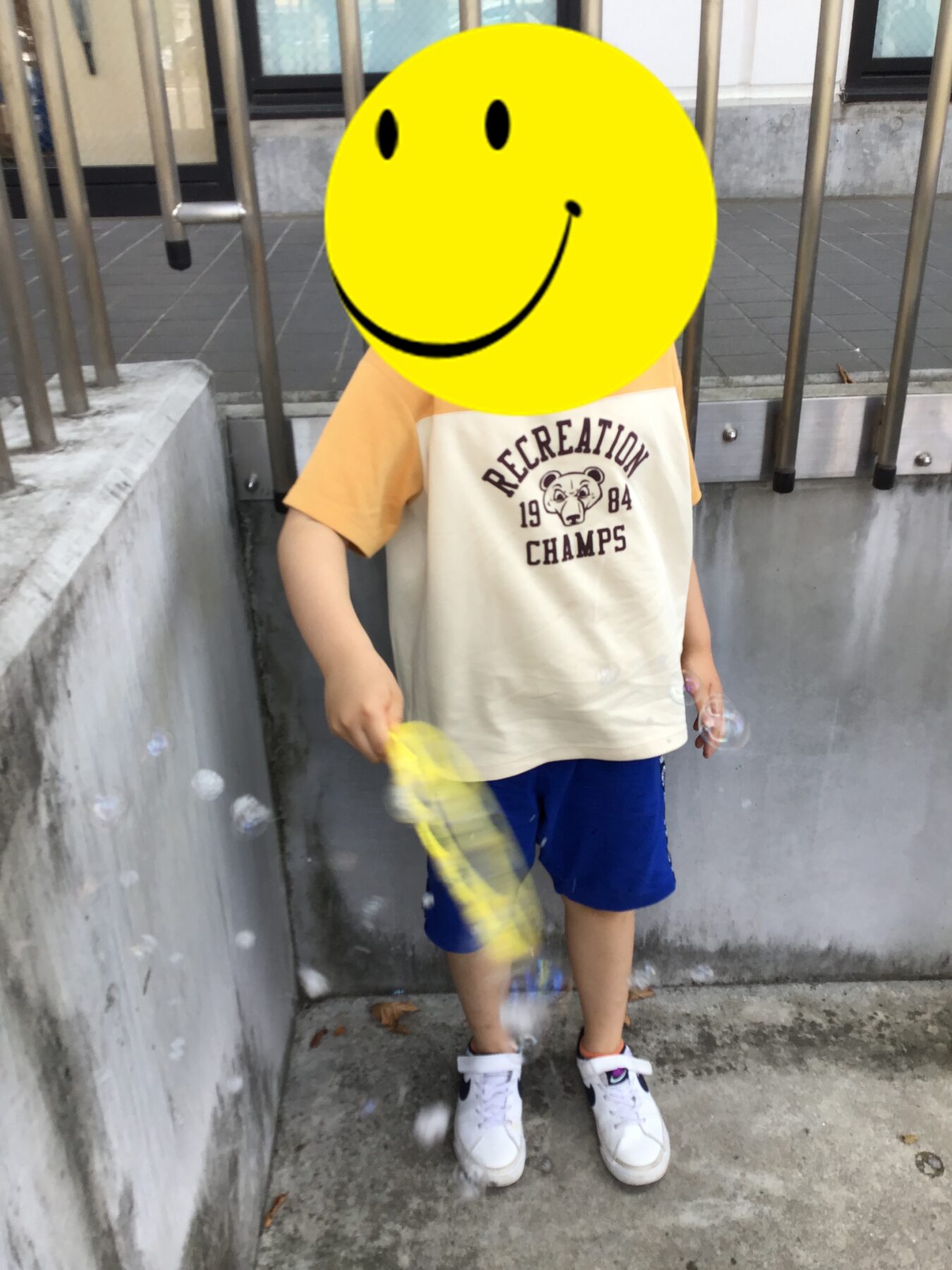 Child standing in a concrete gutter with metal railing, wearing a cream-orange shirt that says 'RECREATION CHAMPS 1984', holding a yellow item.