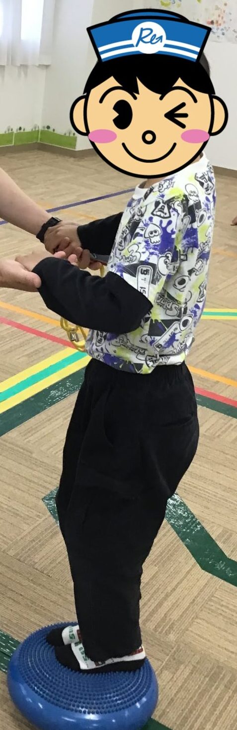 Adult wearing a mask helps a child balance by holding hands during an indoor balance exercise on a blue wobble disc.