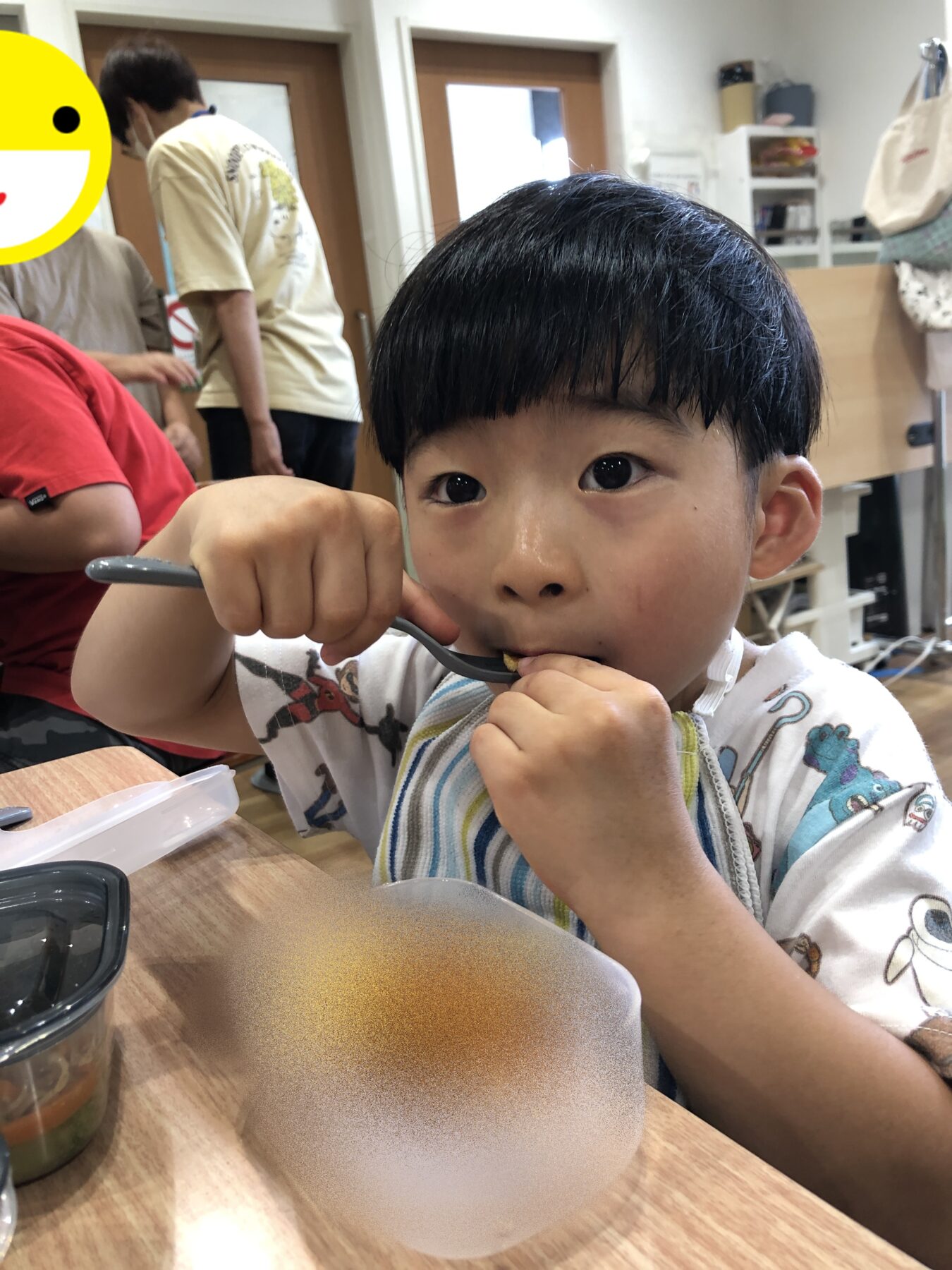 Close-up of a young boy with a bowl haircut eating with a spoon at a table, with adults and a classroom-like setting in the background.