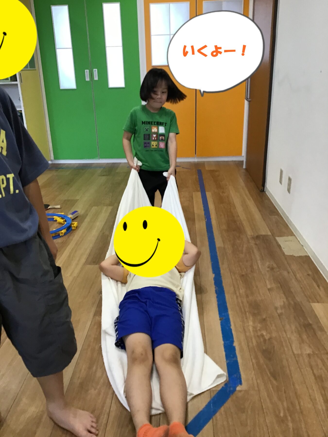 Child in a green t-shirt pushes a white fabric sling with another child lying inside in a gym-like playroom with colorful doors behind them