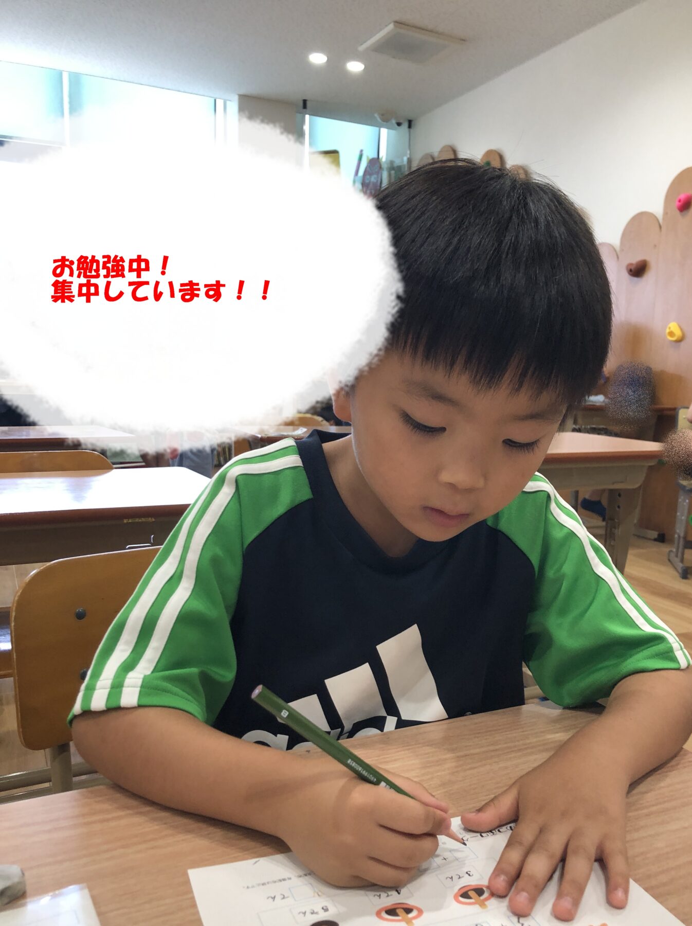 Young boy in a green-sleeved shirt sits at a desk in a classroom, writing on a worksheet with a focused expression. Text overlay in red says he’s studying and concentrating.