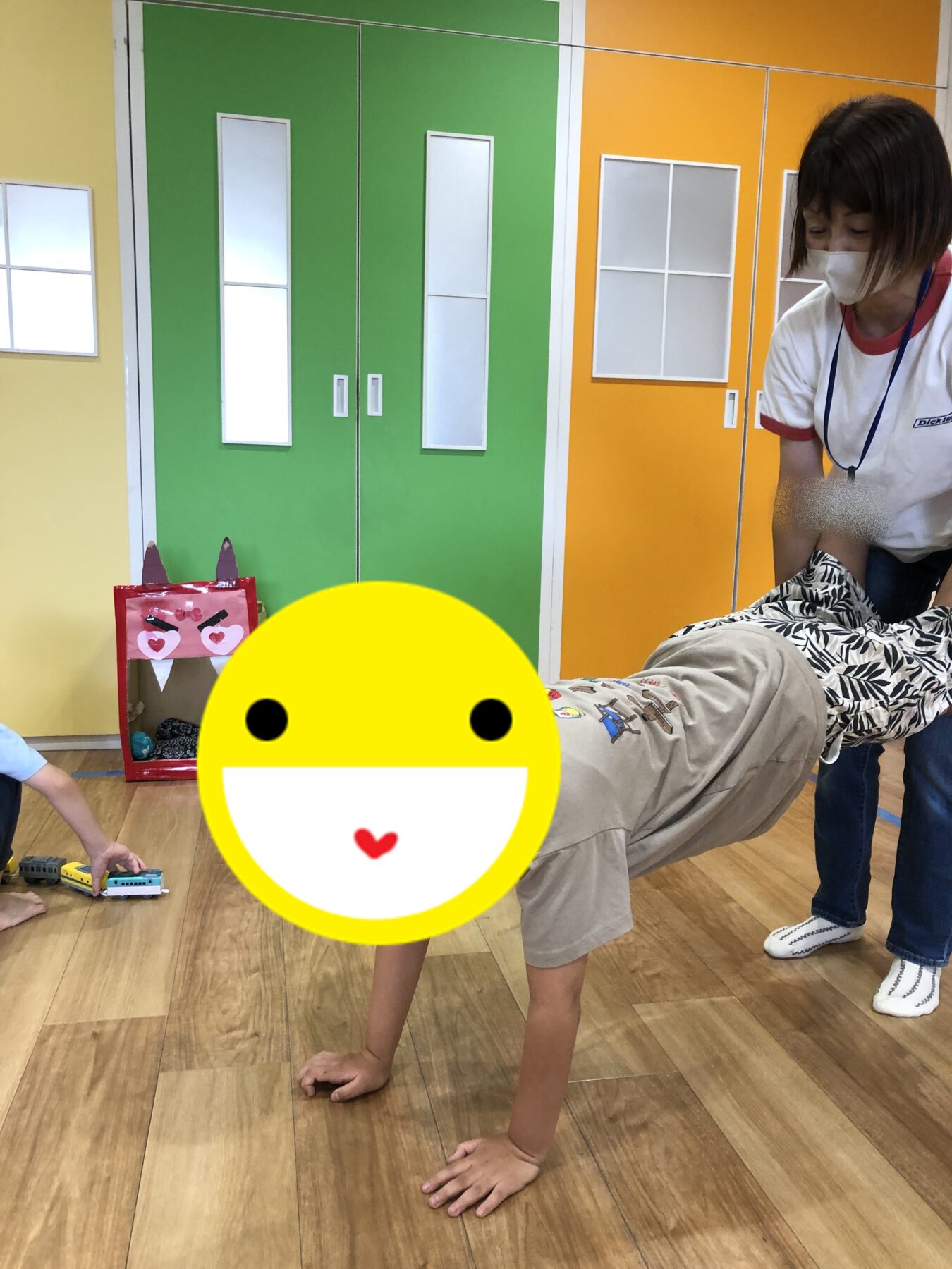 Child on hands and knees crawls on a wooden floor in a bright playroom with green and orange lockers, a masked caregiver nearby.
