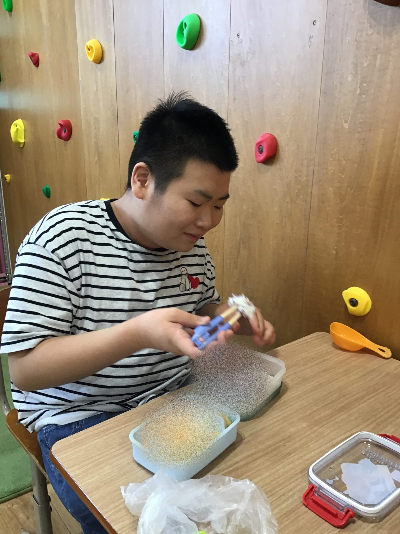 Boy in a striped shirt sits at a table, assembling a craft with blue pieces while sand fills a bin nearby; colorful climbing holds on a wooden wall in the background.