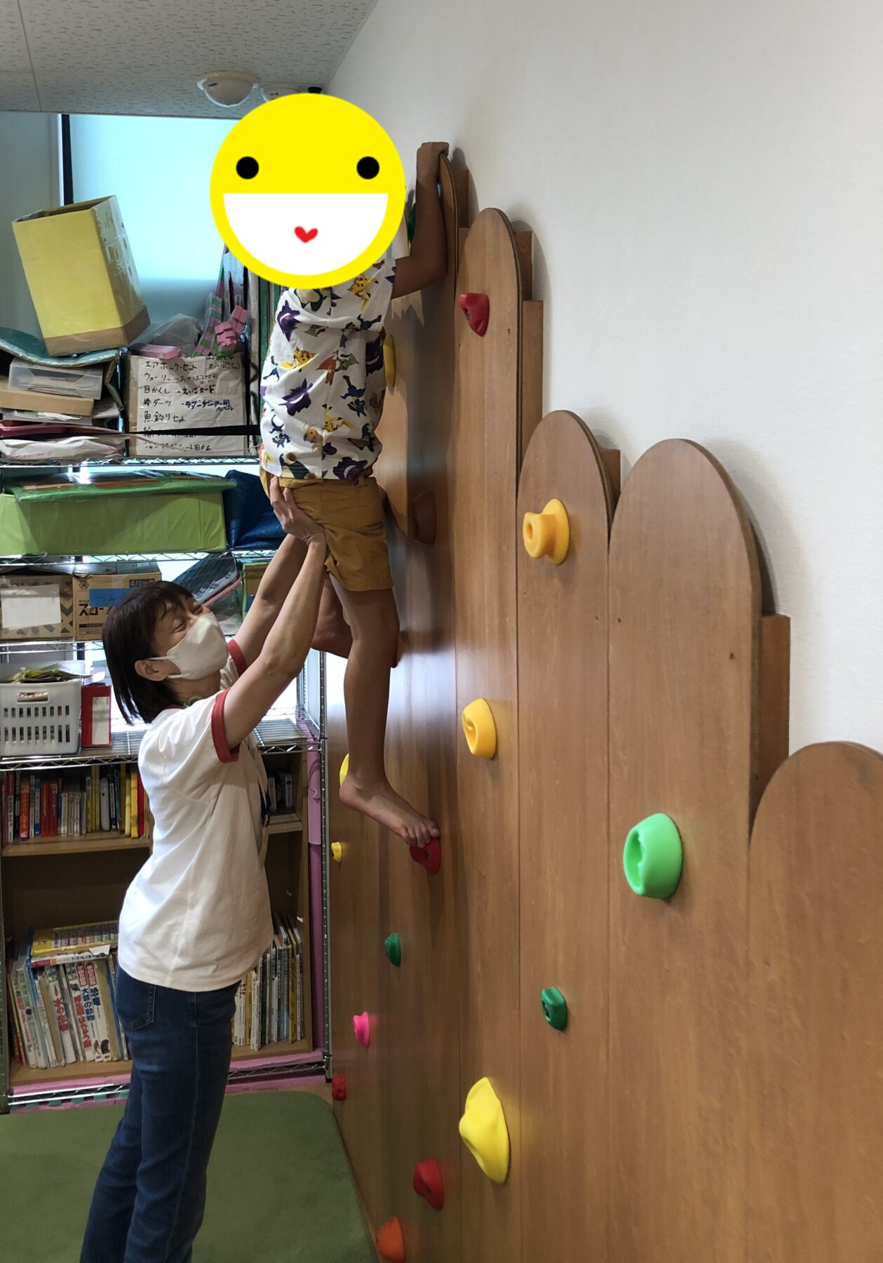 Child wearing a printed shirt and shorts climbs a wooden indoor climbing wall while an adult in a mask steadies them from below.