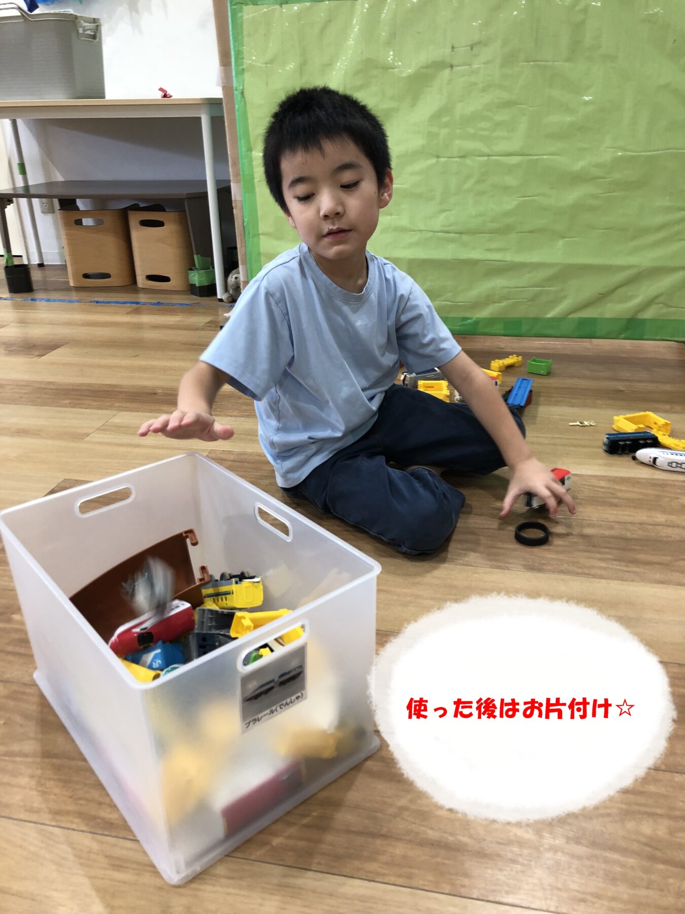 Young boy sits on a wooden floor, playing with colorful plastic building blocks from a clear storage bin.