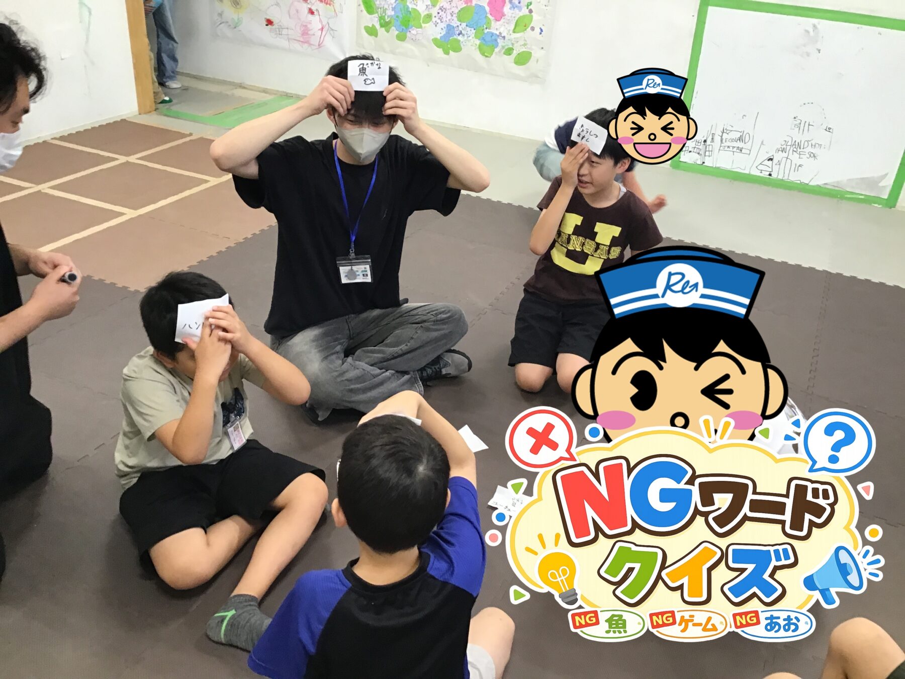 Children and an adult play a classroom game on a soft mat; players hold cards to their foreheads while others watch.