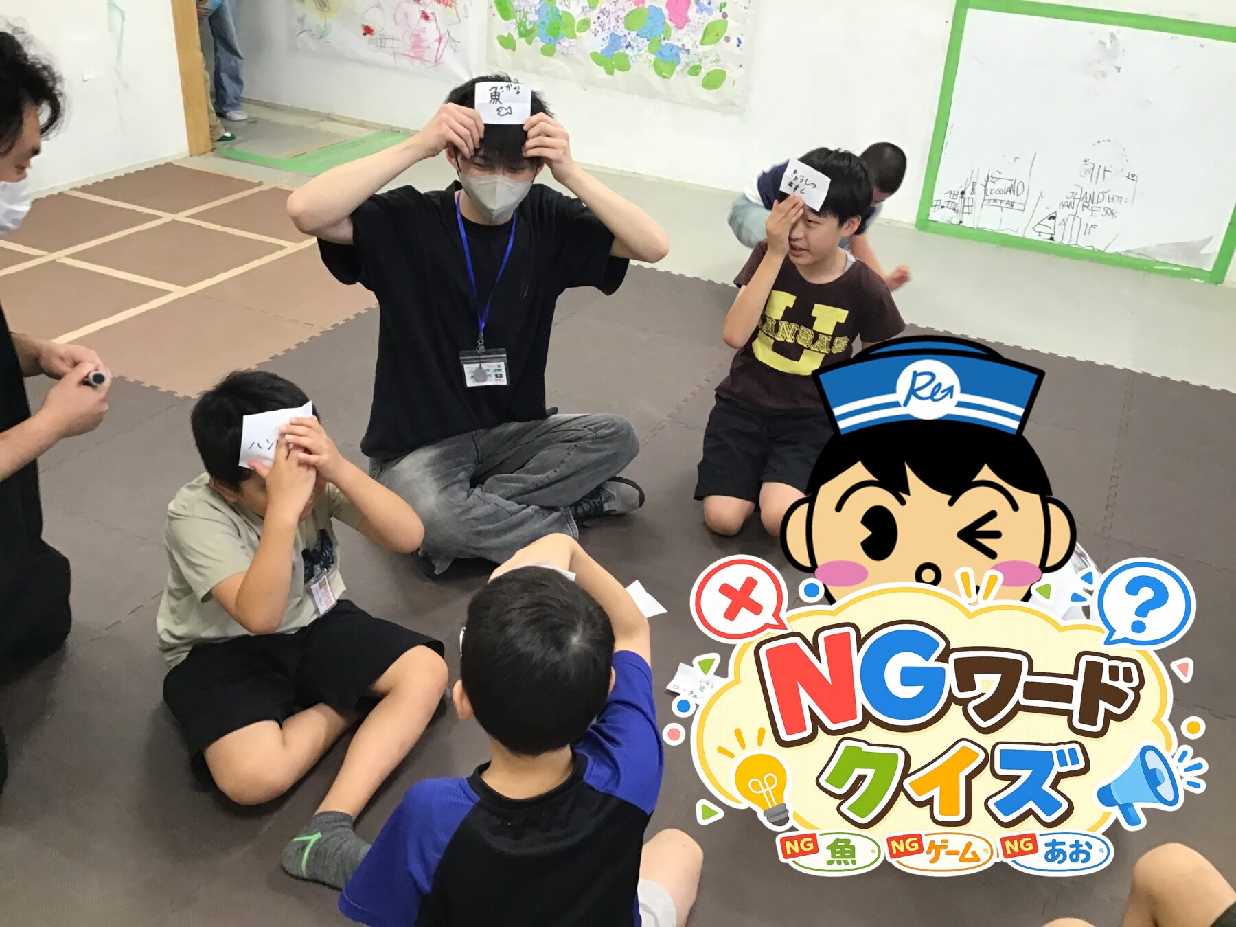 Group of kids sitting on a mats floor playing a guessing game, each with a card on their forehead while an adult supervises.