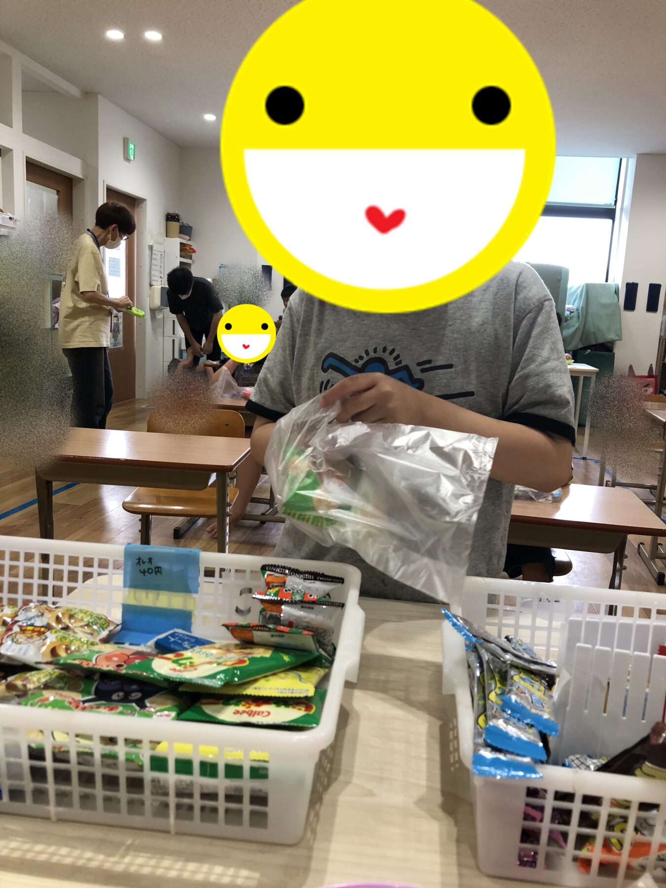 Child standing at a table sorting snack packets in a bright classroom; other kids and an adult in the background help with packaging.