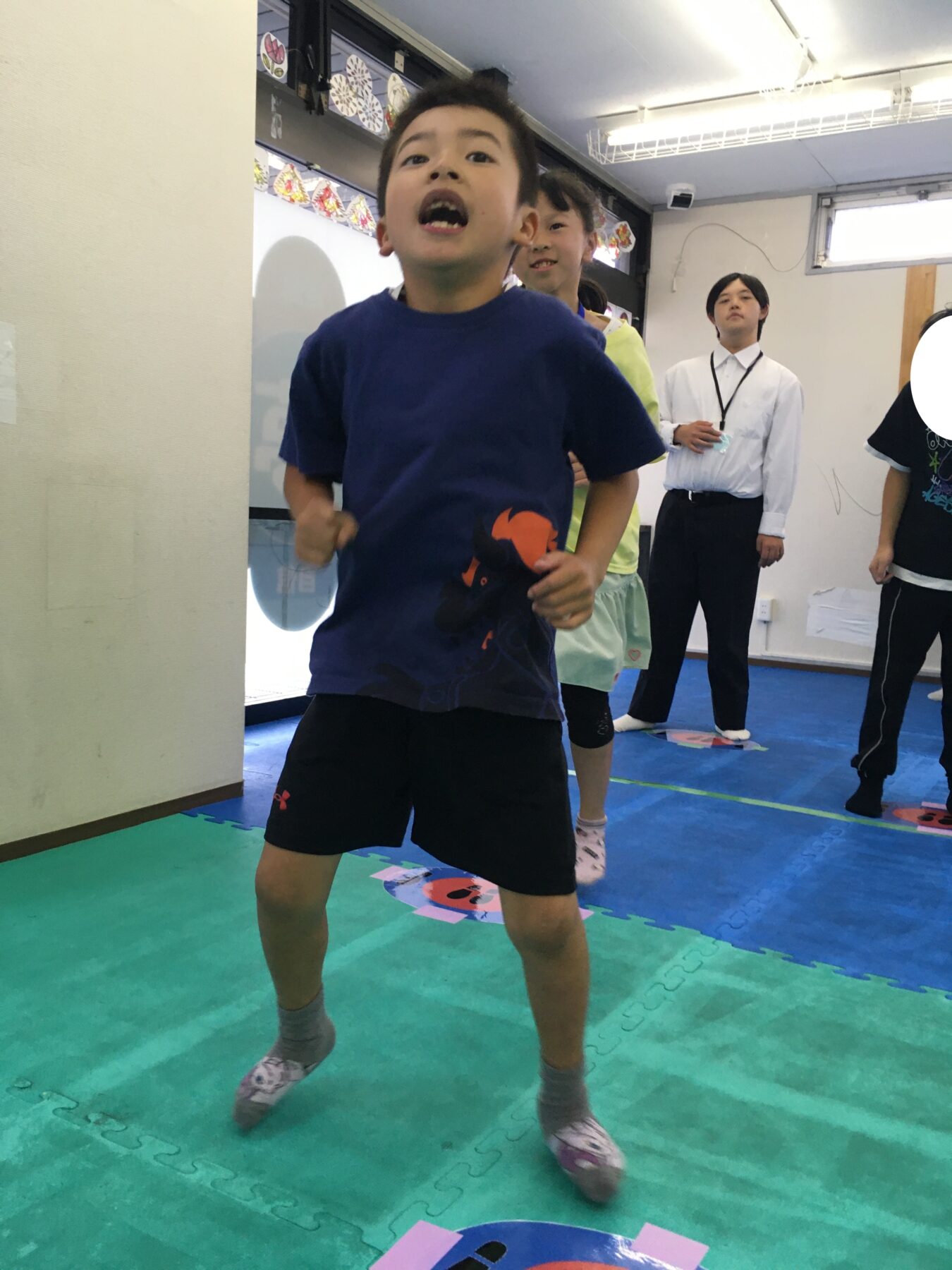 Boy in blue shirt runs toward the camera in a gym-like room with friends and an adult watching in the background.