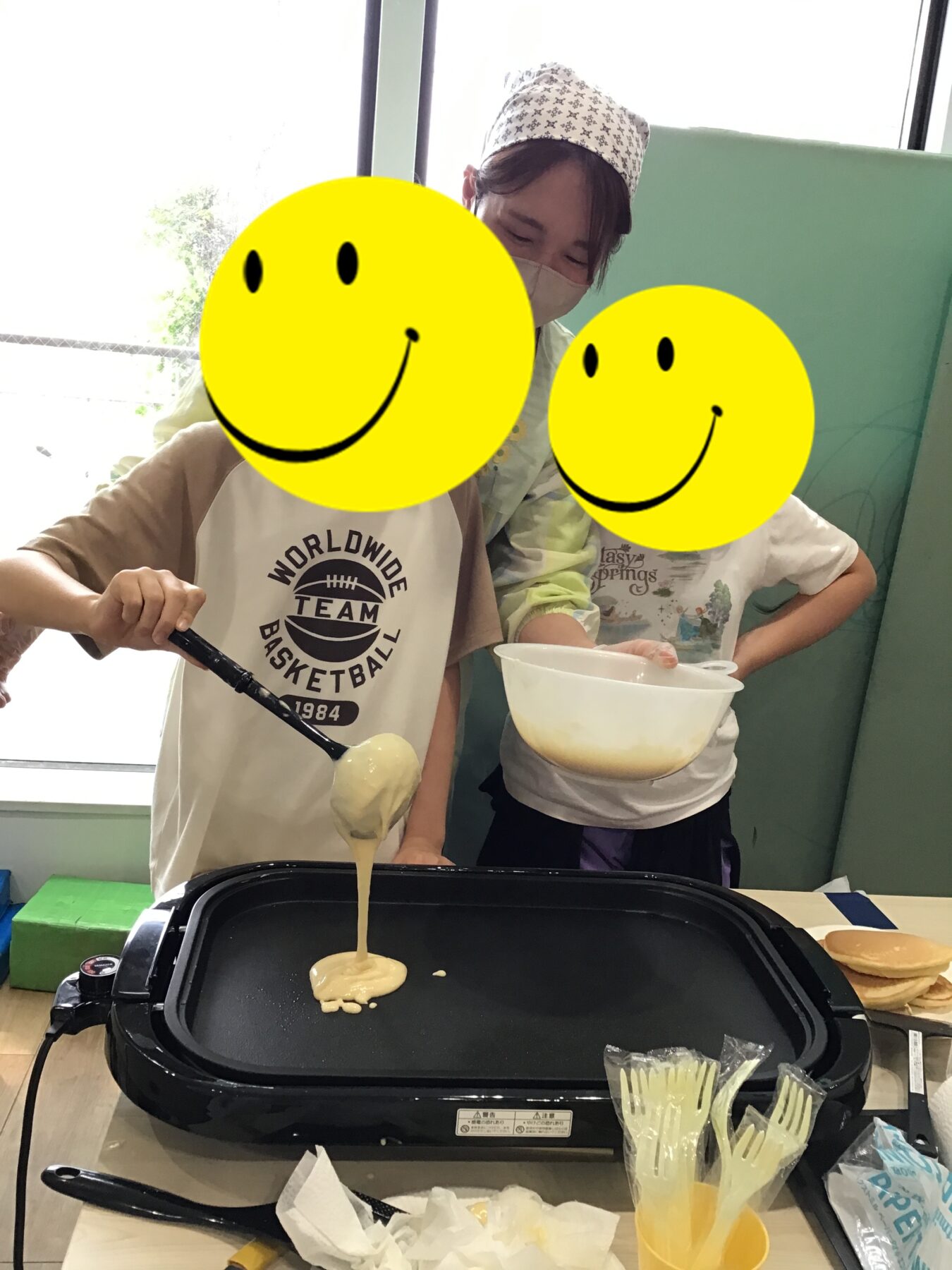 Two kids cooking pancakes on a griddle; one pours batter while the other holds a bowl; faces covered by yellow smiley overlays.