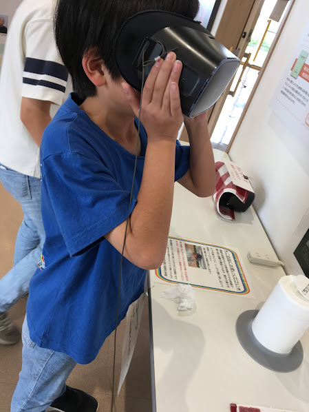Boy in a blue shirt wearing a virtual reality headset at a table, touching the headset with his hands.