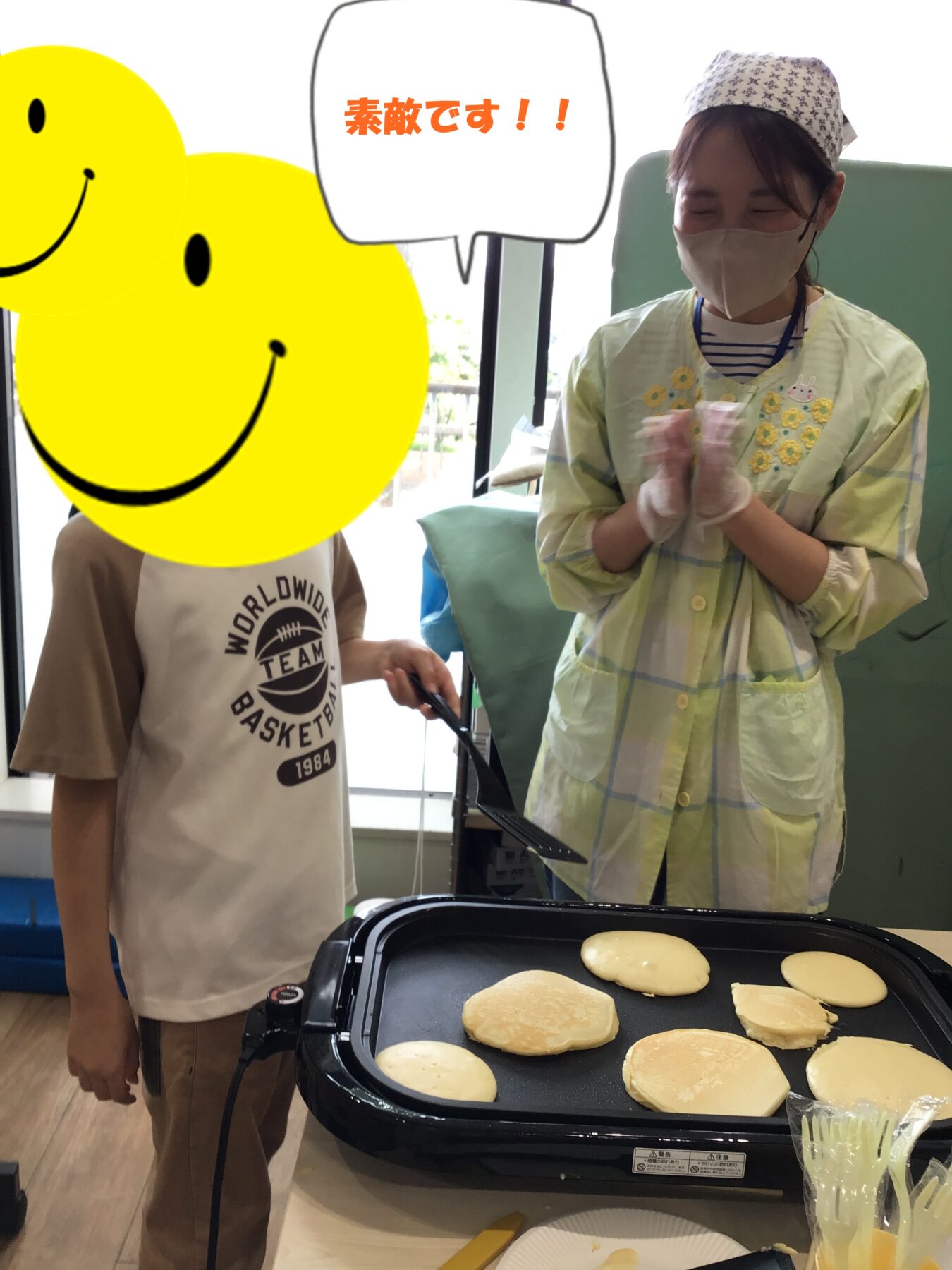 Kitchen scene: pancakes cooking on an electric griddle while a person in a mask and cap claps beside a child whose face is covered by a large yellow smiley overlay; speech bubble reads '素敵です!!'