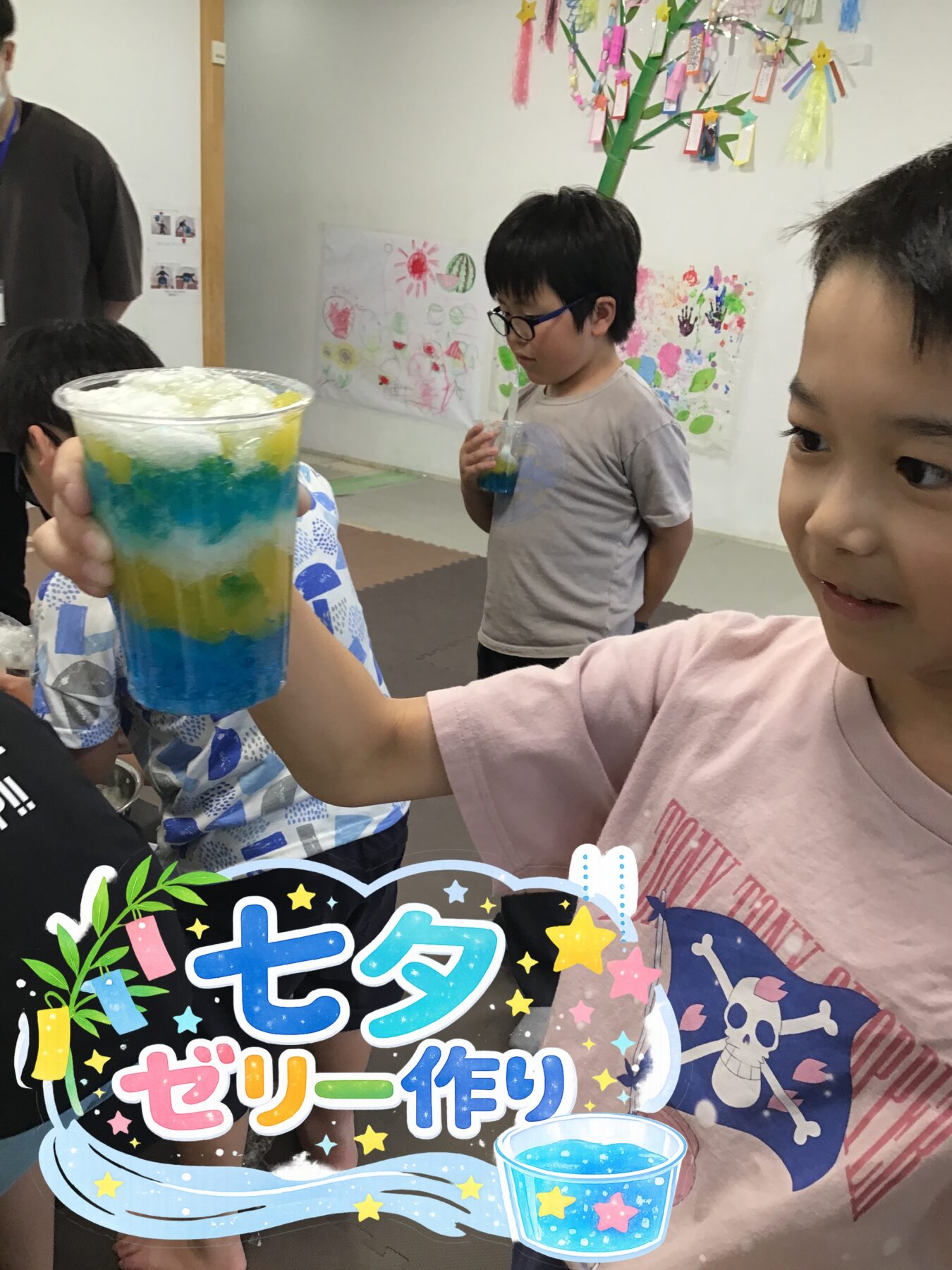 Child proudly shows a tall cup with layered blue, yellow, and white jelly during a kids' jelly-making activity in a classroom.