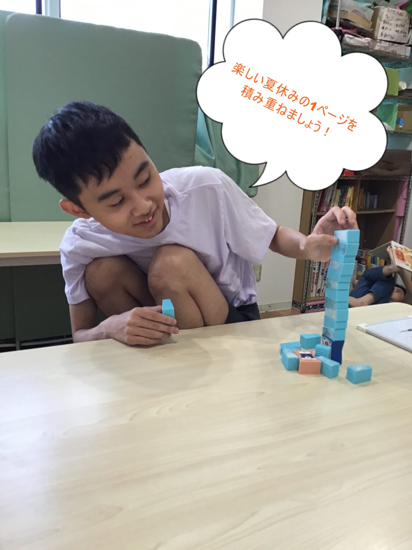 Boy in a white shirt stacking blue building blocks on a table, building a tall tower; bookshelf in background. A speech bubble with orange Japanese text overlays the scene.
