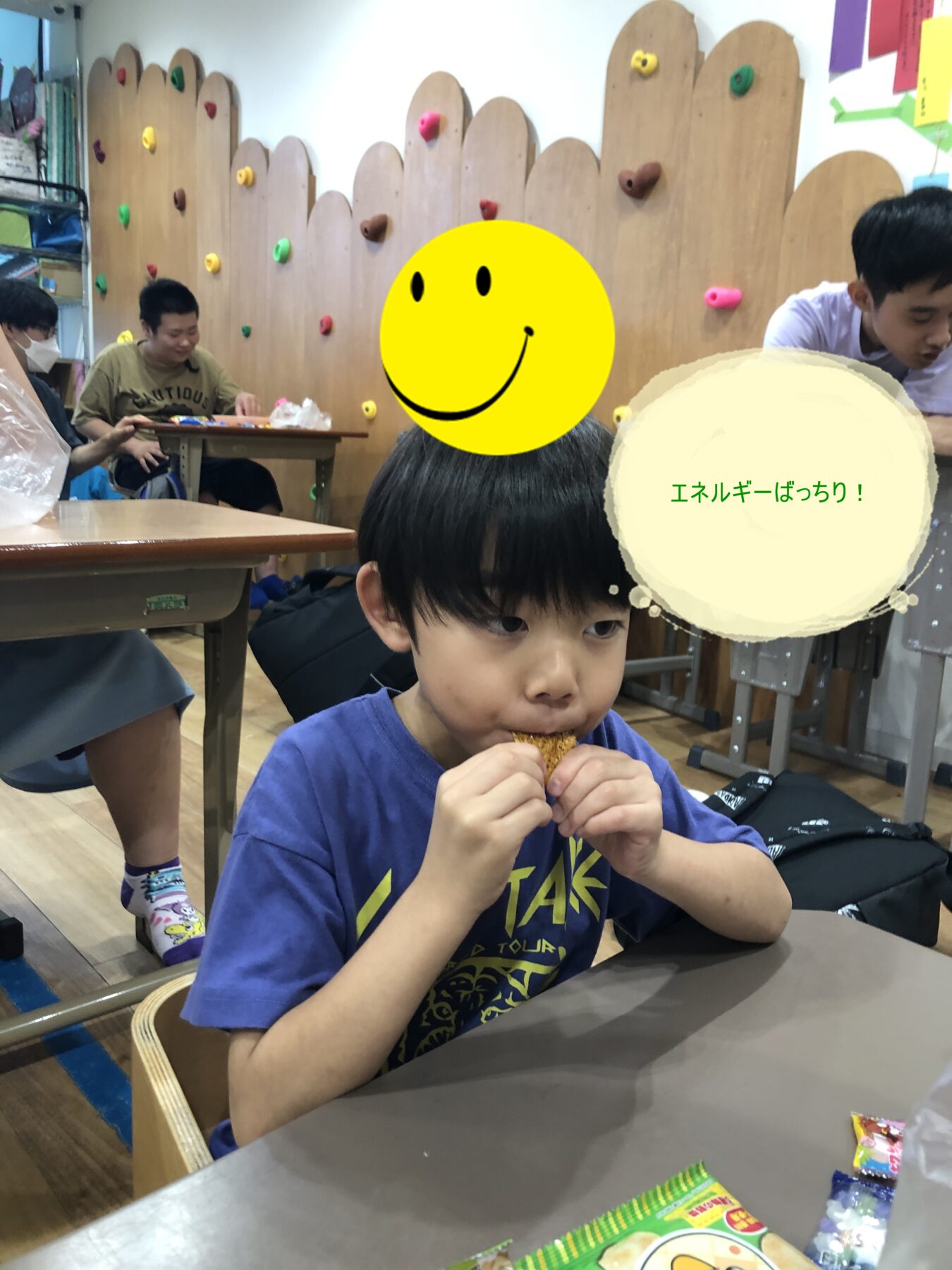 Young boy in a blue shirt eats a snack at a classroom table, with other kids and a colorful climbing-wall backdrop behind him.