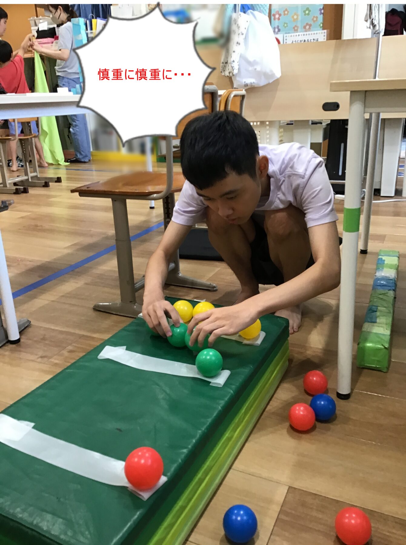 Boy kneeling on a gym mat, placing yellow and green balls on a padded platform; colorful balls scattered nearby and classmates in the background. A speech bubble with red Japanese text reads '慎重に慎重に…'