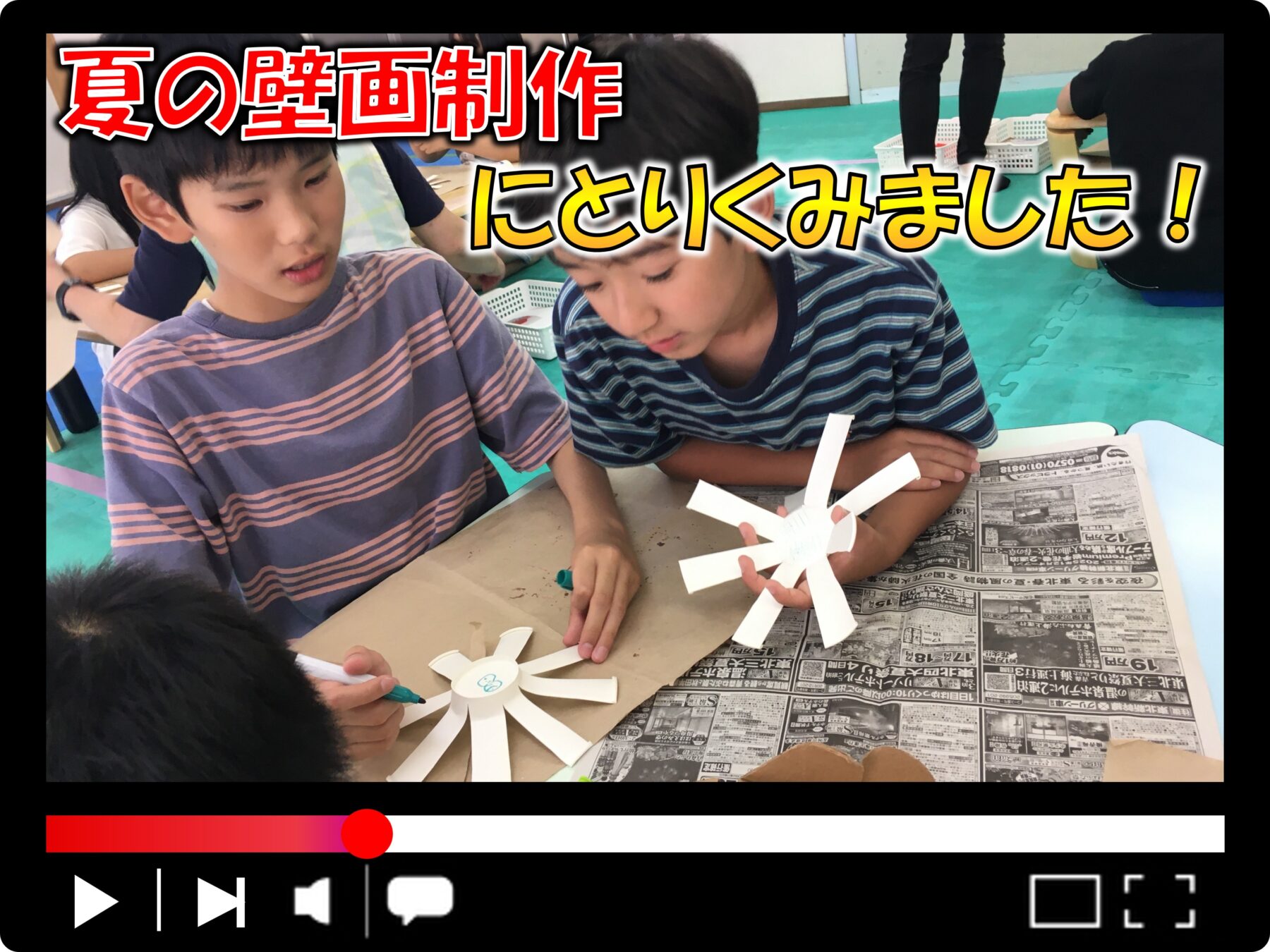 Two children cut white paper into sun shapes for a summer mural, with a newspaper-covered table and a bold Japanese banner overhead reading about summer art.
