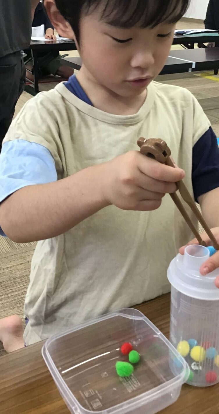 Young child with chopsticks scooping chocolate over a cup at a craft table, colorful pom-poms in foreground, adults seated in background.