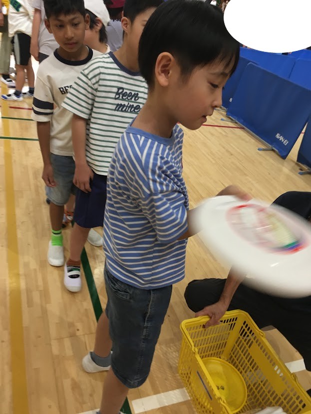 Boy in a blue striped shirt hands a white frisbee to an adult near a yellow basket in a gym, others line up behind him.