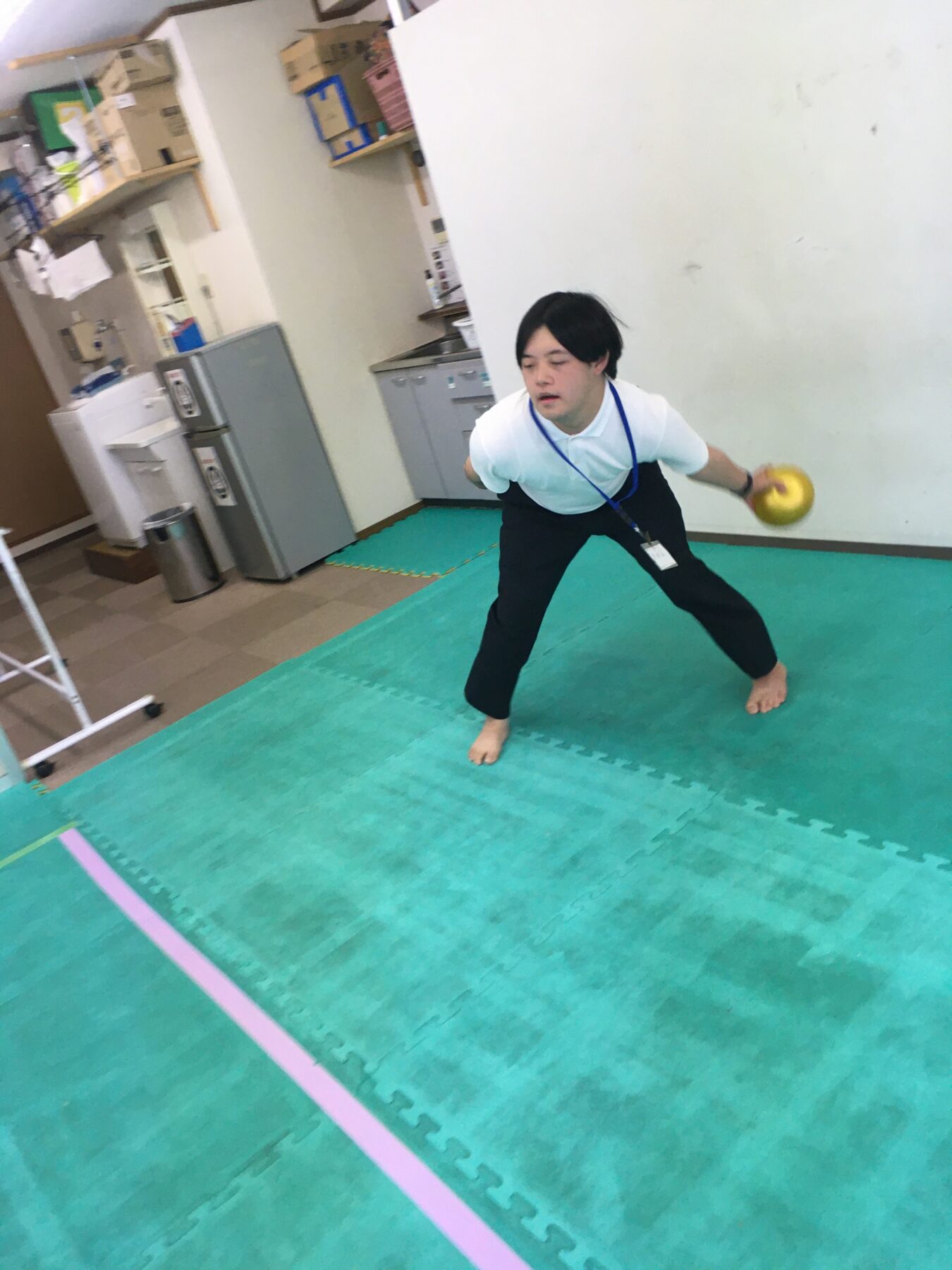 Man in a white polo shirt and black pants, barefoot, lunging with a yellow kettlebell in his right hand on green mats.