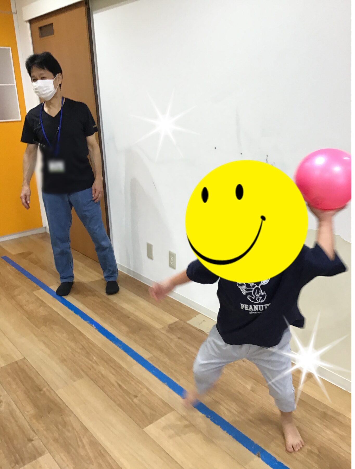 Man in a mask and lanyard watches as a child with a yellow smiley sticker on their face throws a pink ball in a gym.