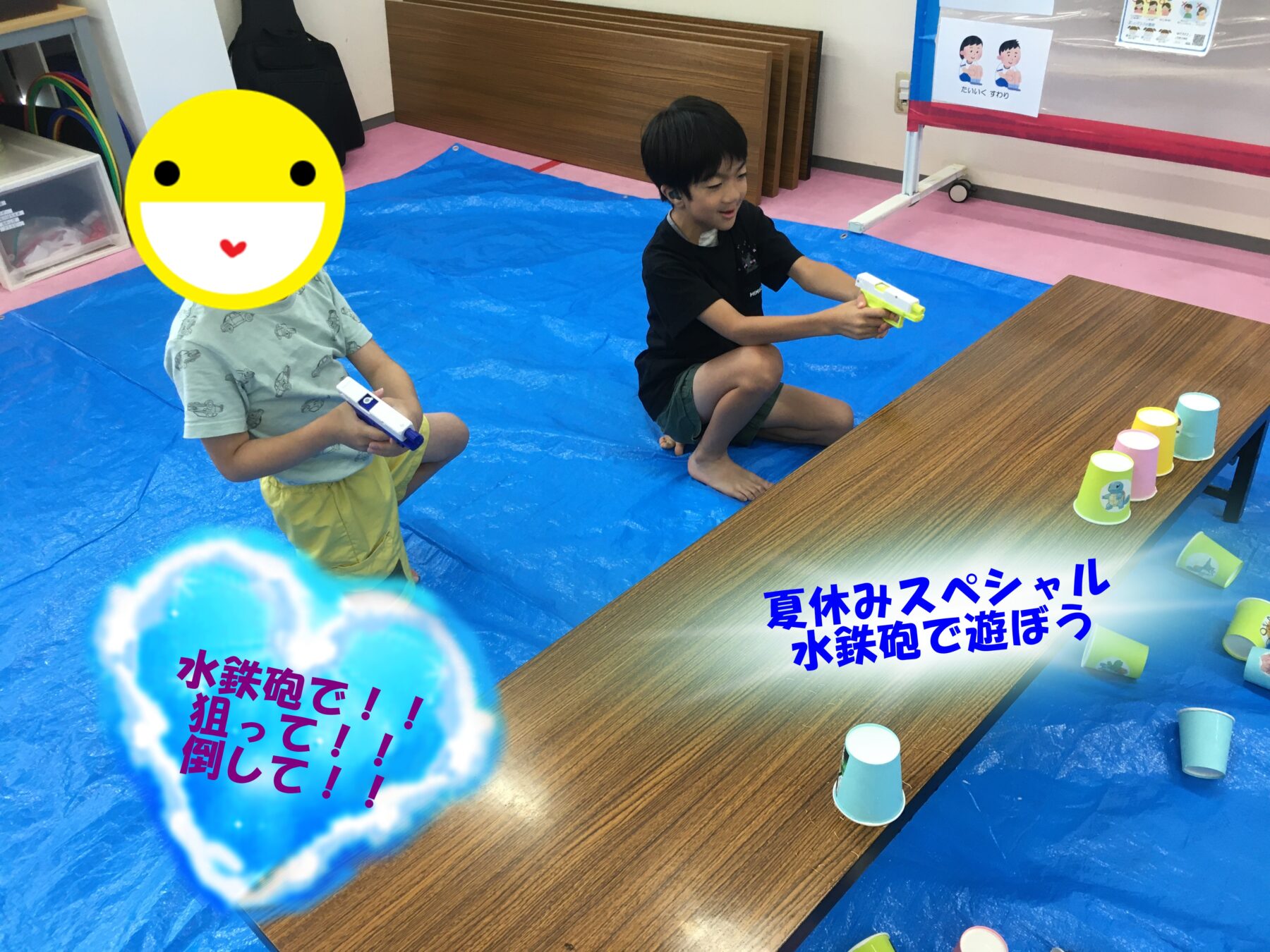 Two children kneeling on a blue mat, each holding a water gun in a playful indoor water game area with cups on a table nearby.