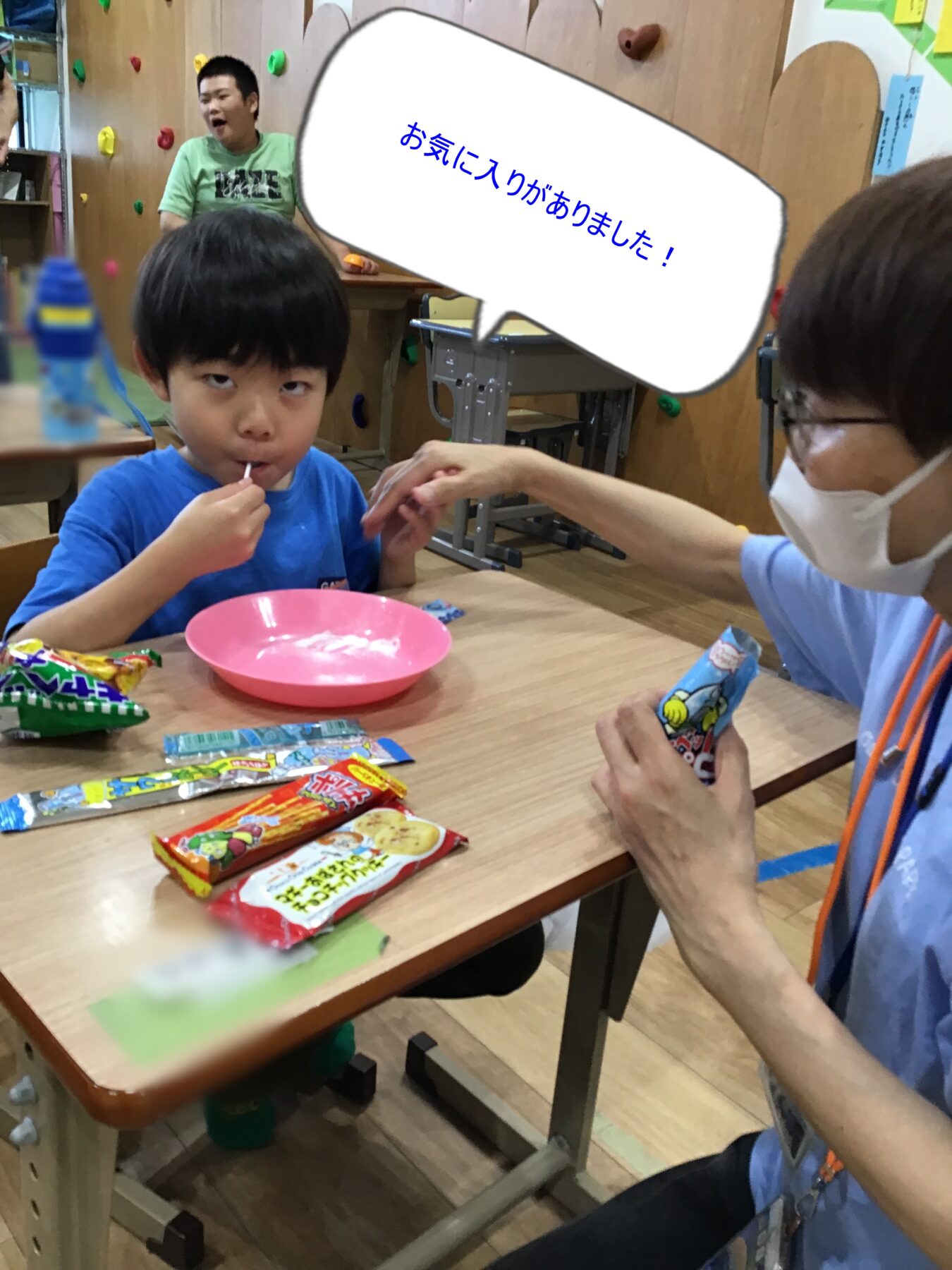 Young boy in a blue shirt sits at a table and is fed a snack by a masked adult; colorful candy wrappers and a pink bowl on the table behind him.