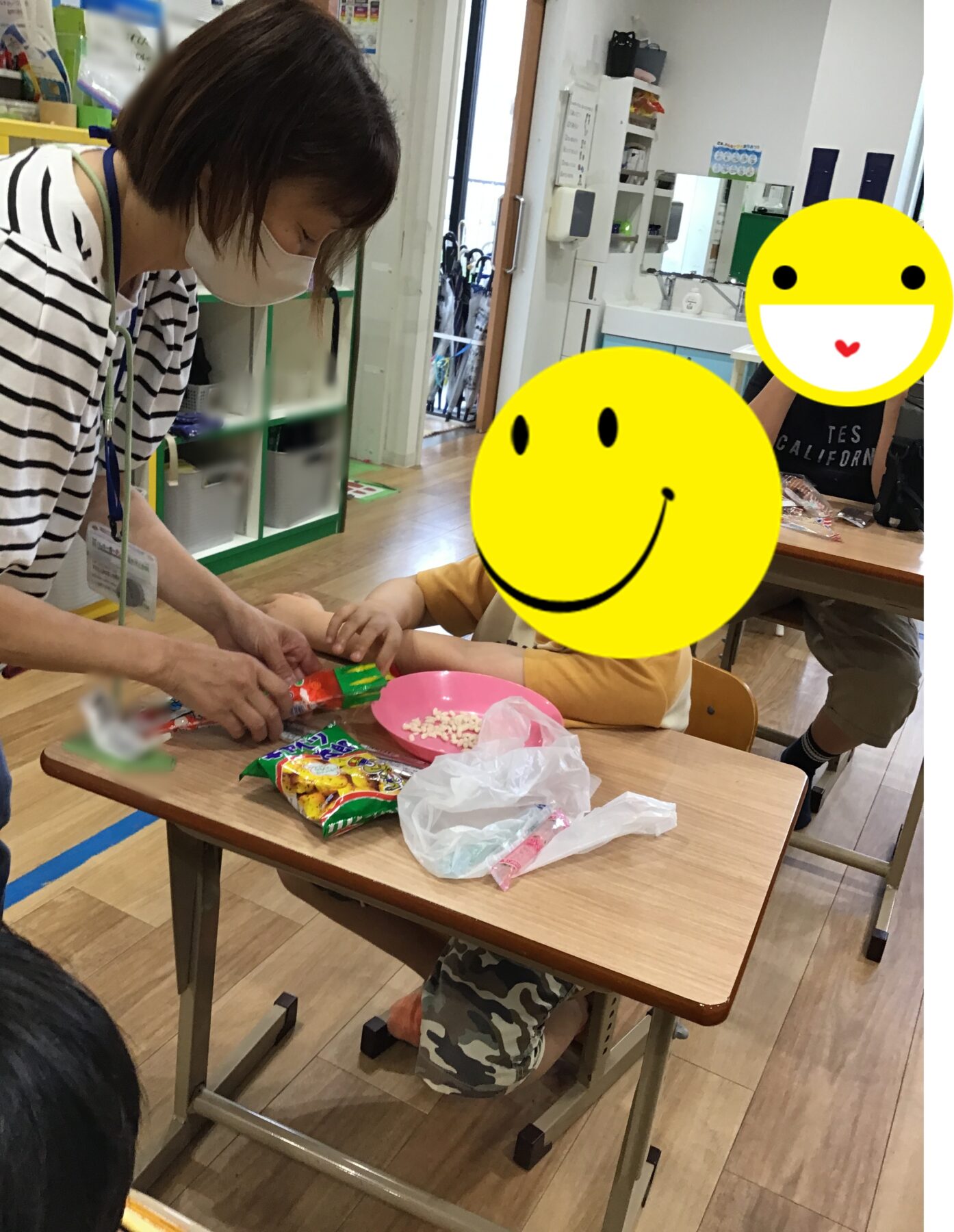 Teacher in a striped shirt and mask helps a child unwrap candy at a classroom table with snack bags and a pink bowl nearby.