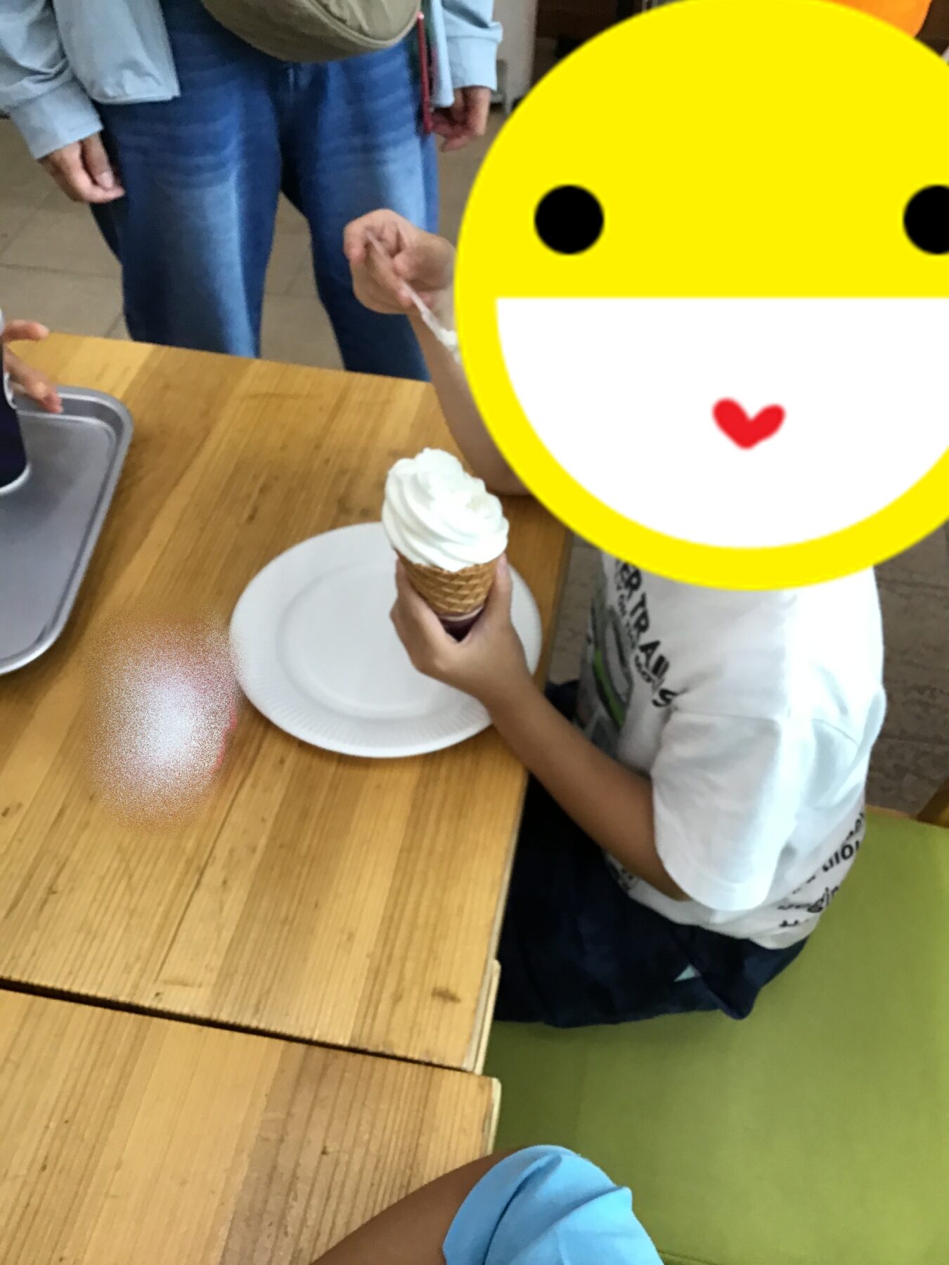 Child holding an ice cream cone with whipped cream at a wooden table; a bright yellow smiley overlay covers part of the scene.