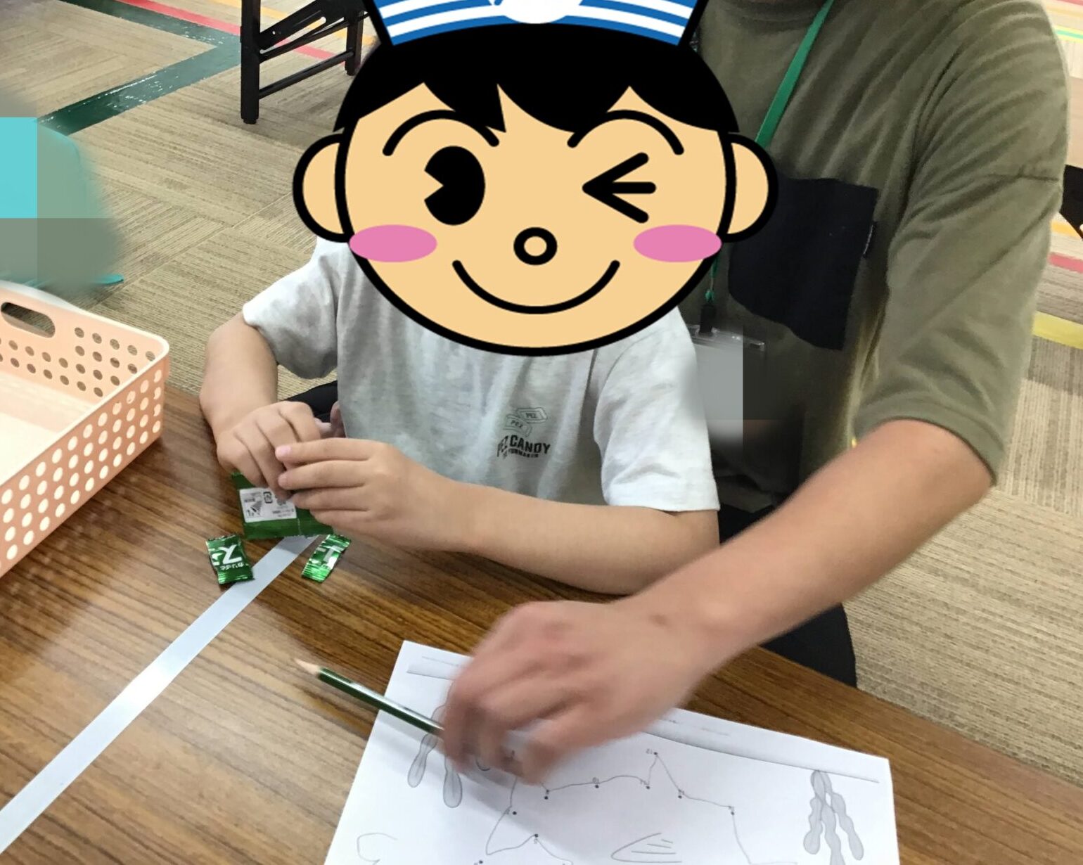 Adult assisting a child at a wooden table with a pencil and paper; colorful snack wrappers and a pink basket nearby.