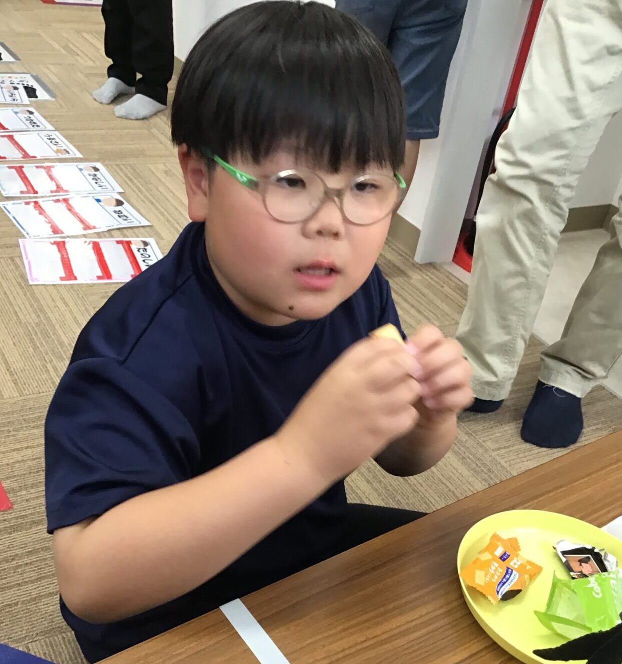Young boy with green glasses eating a snack at a table, with a yellow plate of wrappers nearby.
