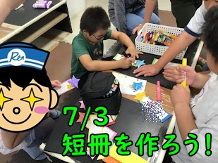 Children in a classroom around a table doing a craft activity with colorful paper pieces; a cartoon sticker is on the left and green Japanese text overlay appears across the image.