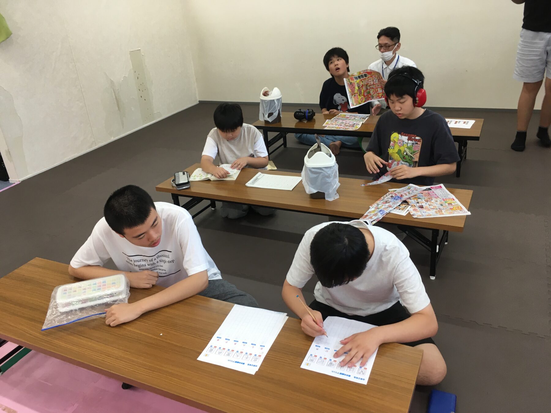 Several youths sit at long desks, working on worksheets in a classroom; one wears red earmuffs, others focus on papers with colorful magazines nearby.