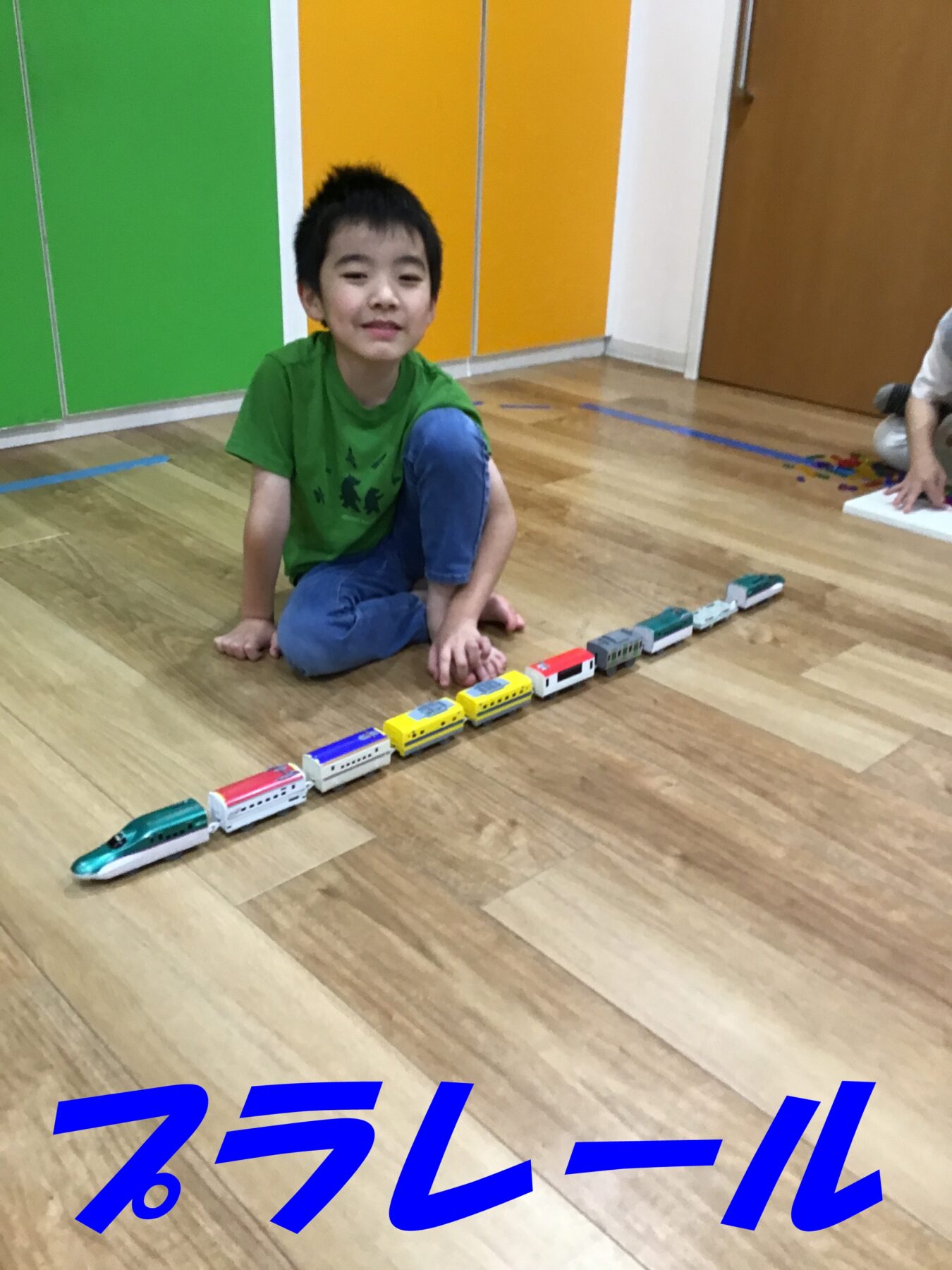 Young boy in a green shirt sits on a wooden floor, playing with a colorful toy train set lined up in front of him.