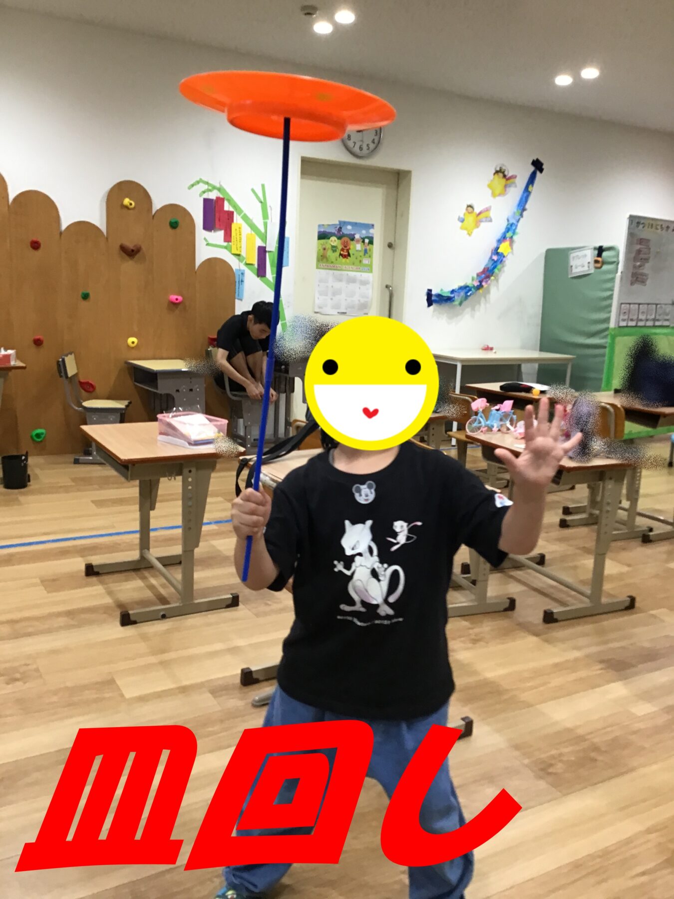 Child in a classroom holding a tall blue pole with an orange circular disc on top, performing a balancing toy activity; colorful wall decorations and desks in the background; large red text overlay partially visible at the bottom.