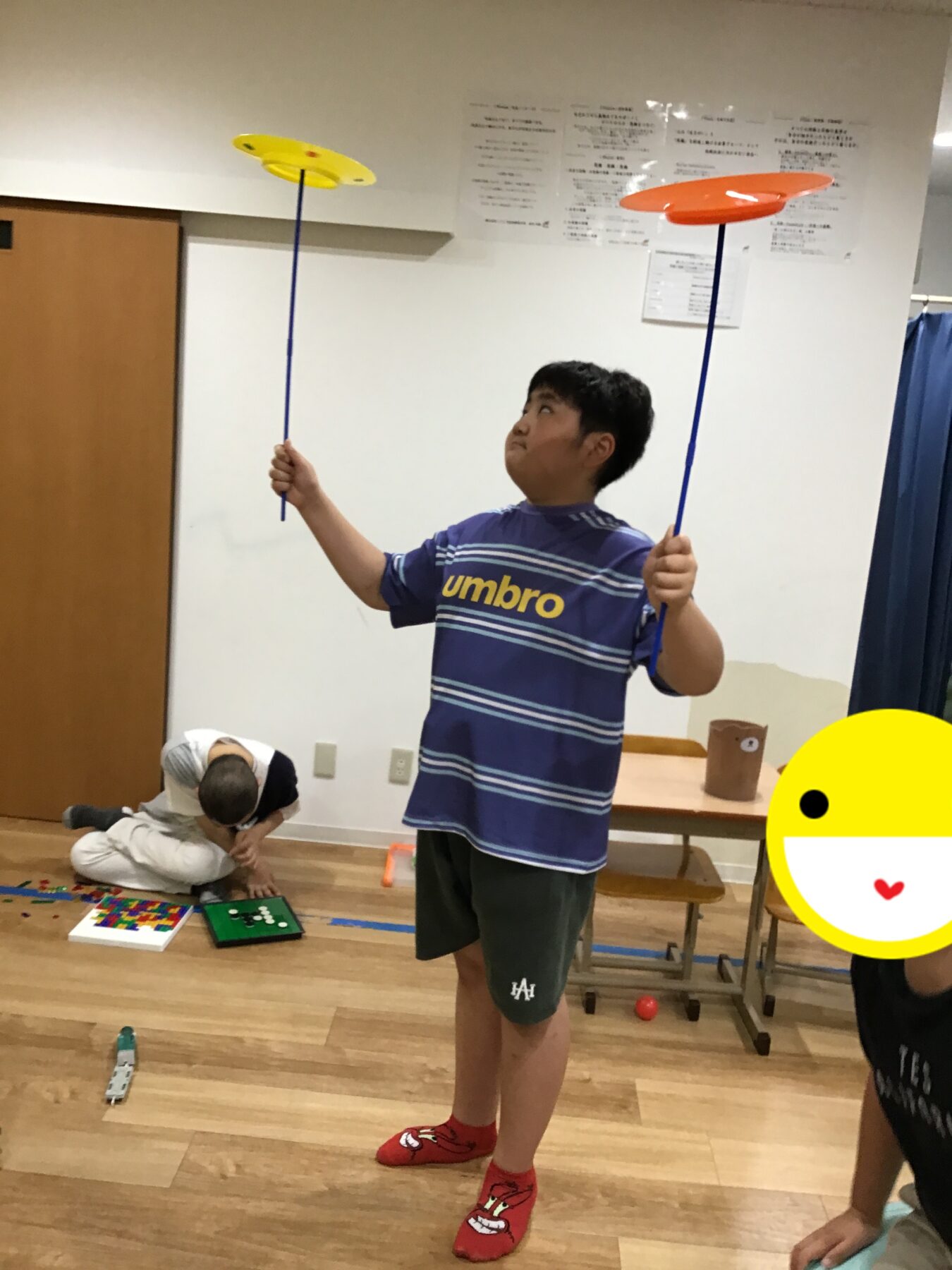 Boy in a blue striped shirt balances yellow and orange plates on long blue sticks indoors, with another child playing on the floor nearby.
