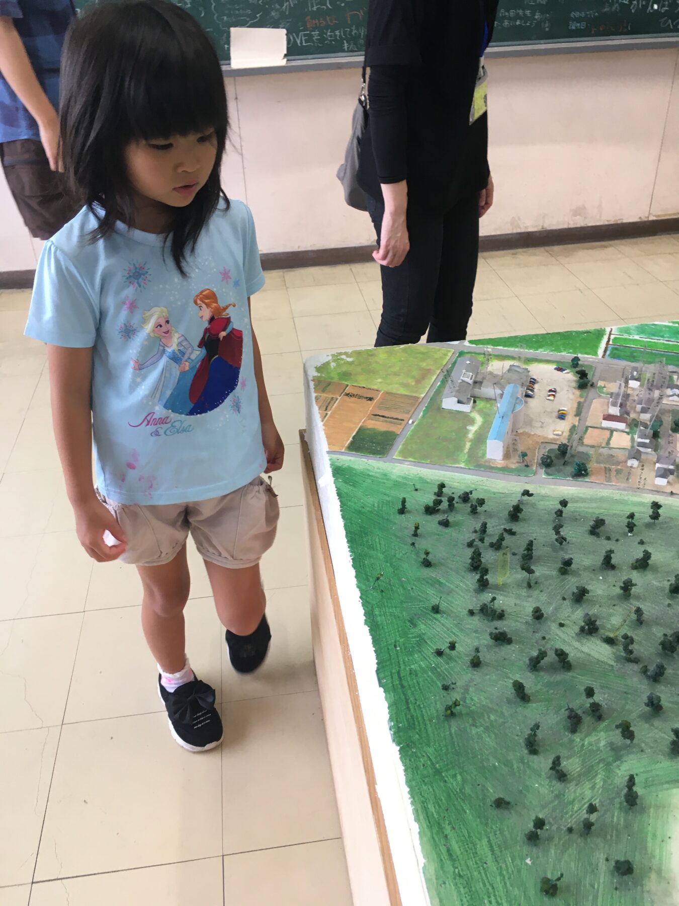 Young girl in a blue Elsa shirt examining a large tabletop model of a town.