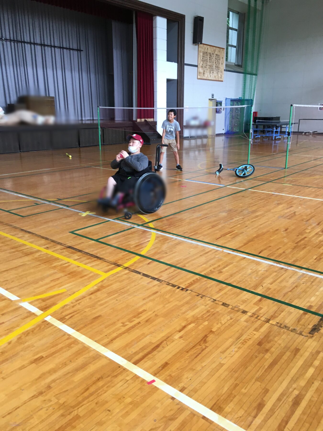Boy stands by a badminton net in a gym as a man in a wheelchair holds a ball nearby.