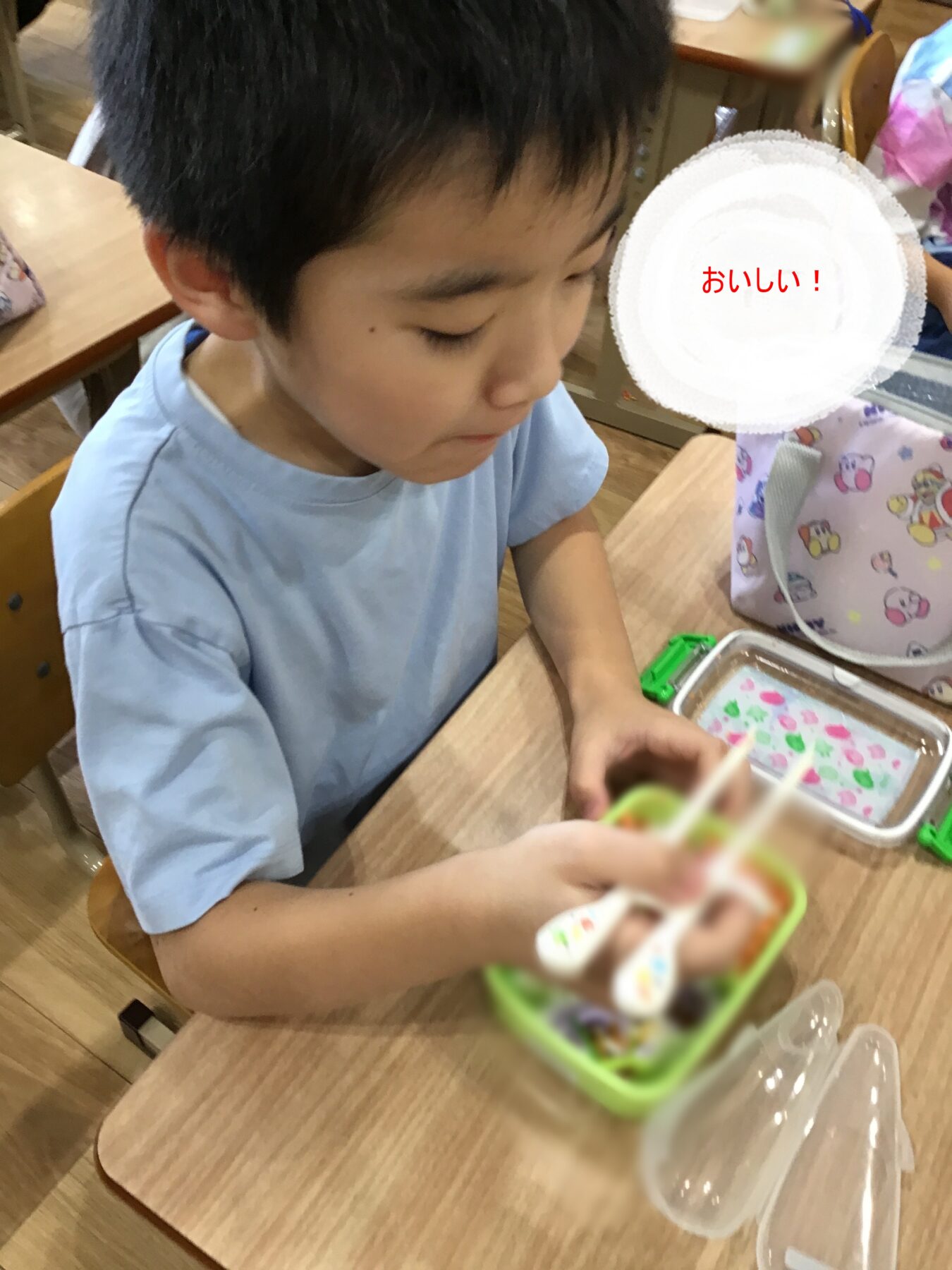 Young boy in a light blue shirt eating lunch at a school desk, using chopsticks from a green lunch box.