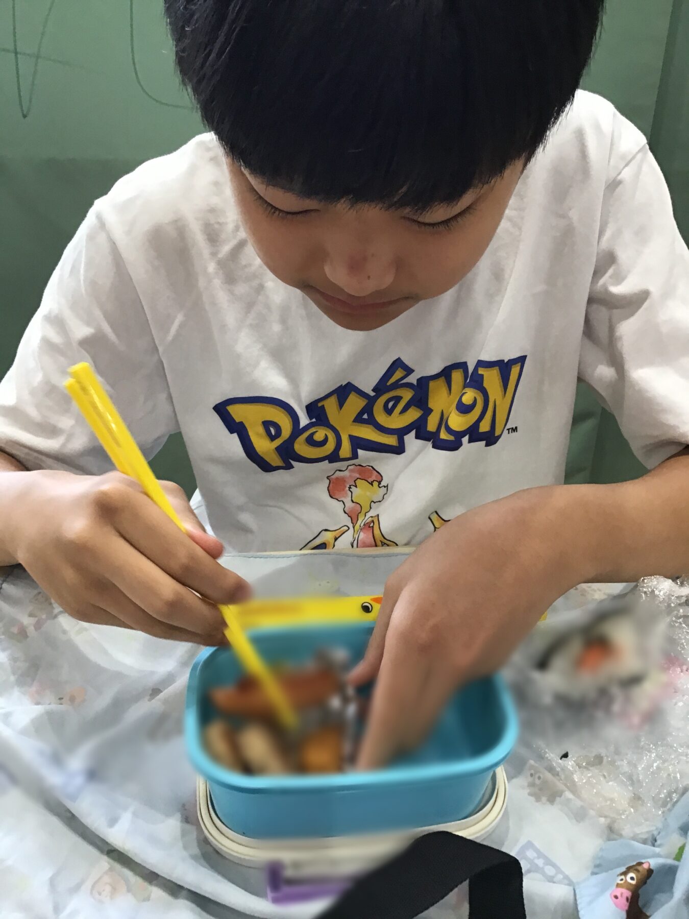 Child drawing with a yellow pencil, wearing a white Pokemon shirt, at a table with a blue lunchbox nearby (top view).