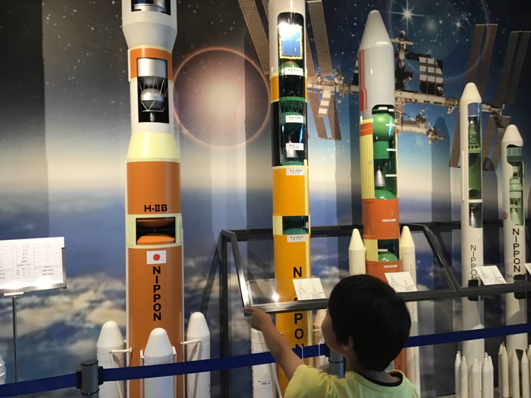 Child in a yellow shirt points at a tall orange-and-white rocket model in a space museum display, with other rockets in the background.