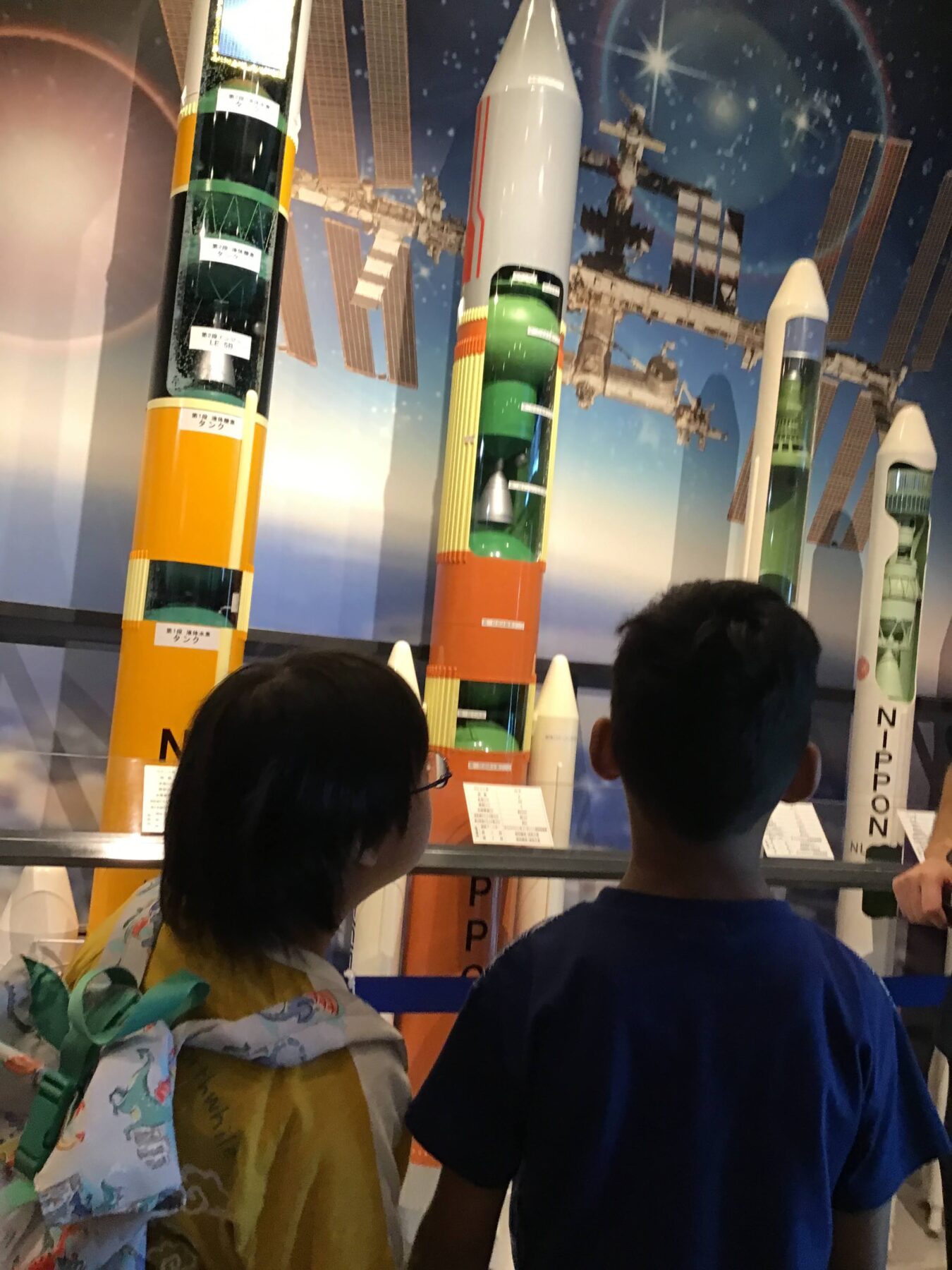 Two children stand at a museum display of tall rocket models against a space-themed backdrop, watching the exhibits curiously.