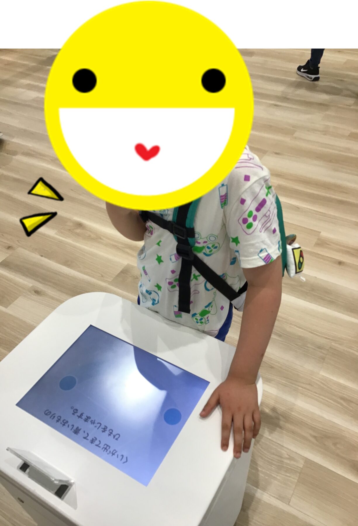 Child with a yellow smiley sticker over their face, standing at a white desk with a blue worksheet on it in a room with a wood floor