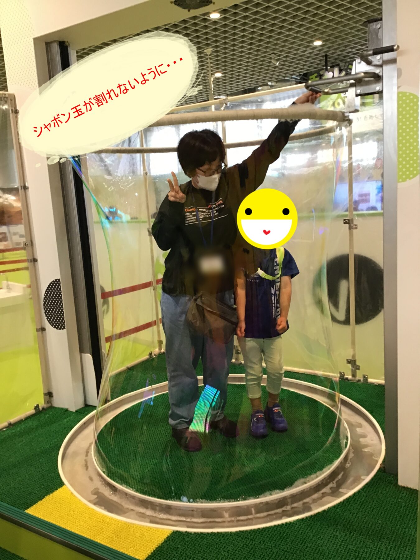 Adult and child posing inside a transparent bubble enclosure at a science/interactive exhibit; a speech bubble above reads in Japanese 'Shabontama ga ware nai you ni…'