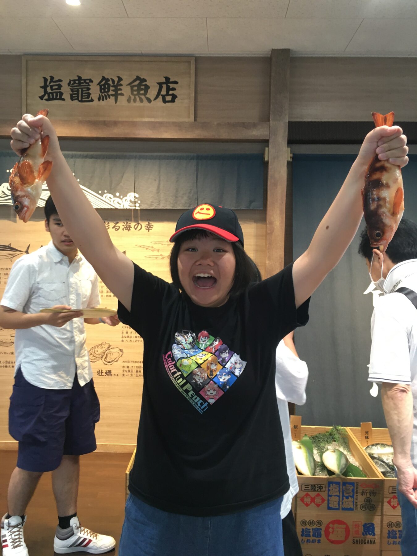 Woman wearing a black T-shirt and red cap raises two fish by their tails in a seafood market, smiling at the camera with seafood crates in the background.