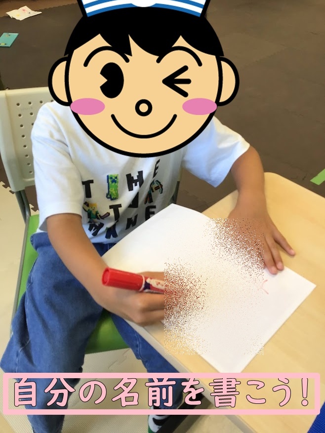 Child at a desk writing their name with a red marker; bottom banner reads 自分の名前を書こう!