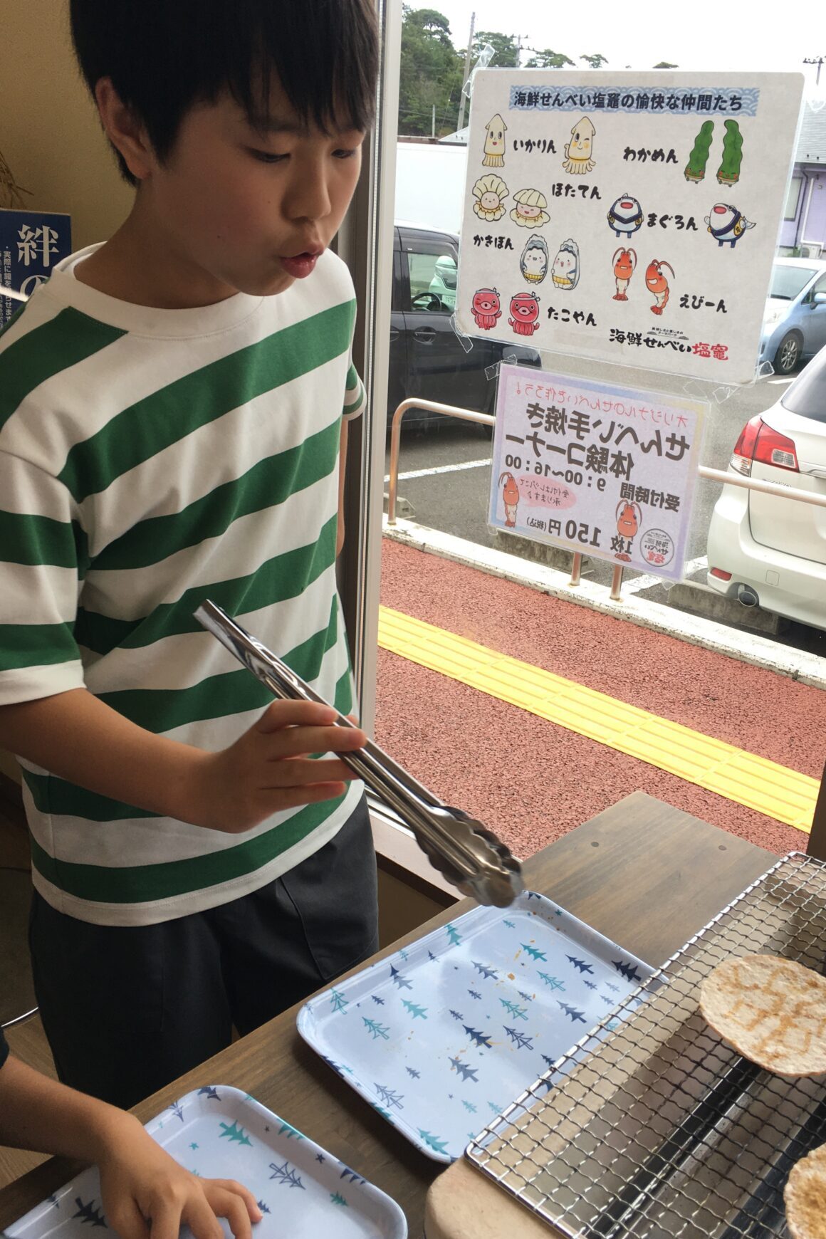 Boy in a green-and-white striped shirt uses tongs to grill bread slices at a counter, posters visible outside the window behind him.