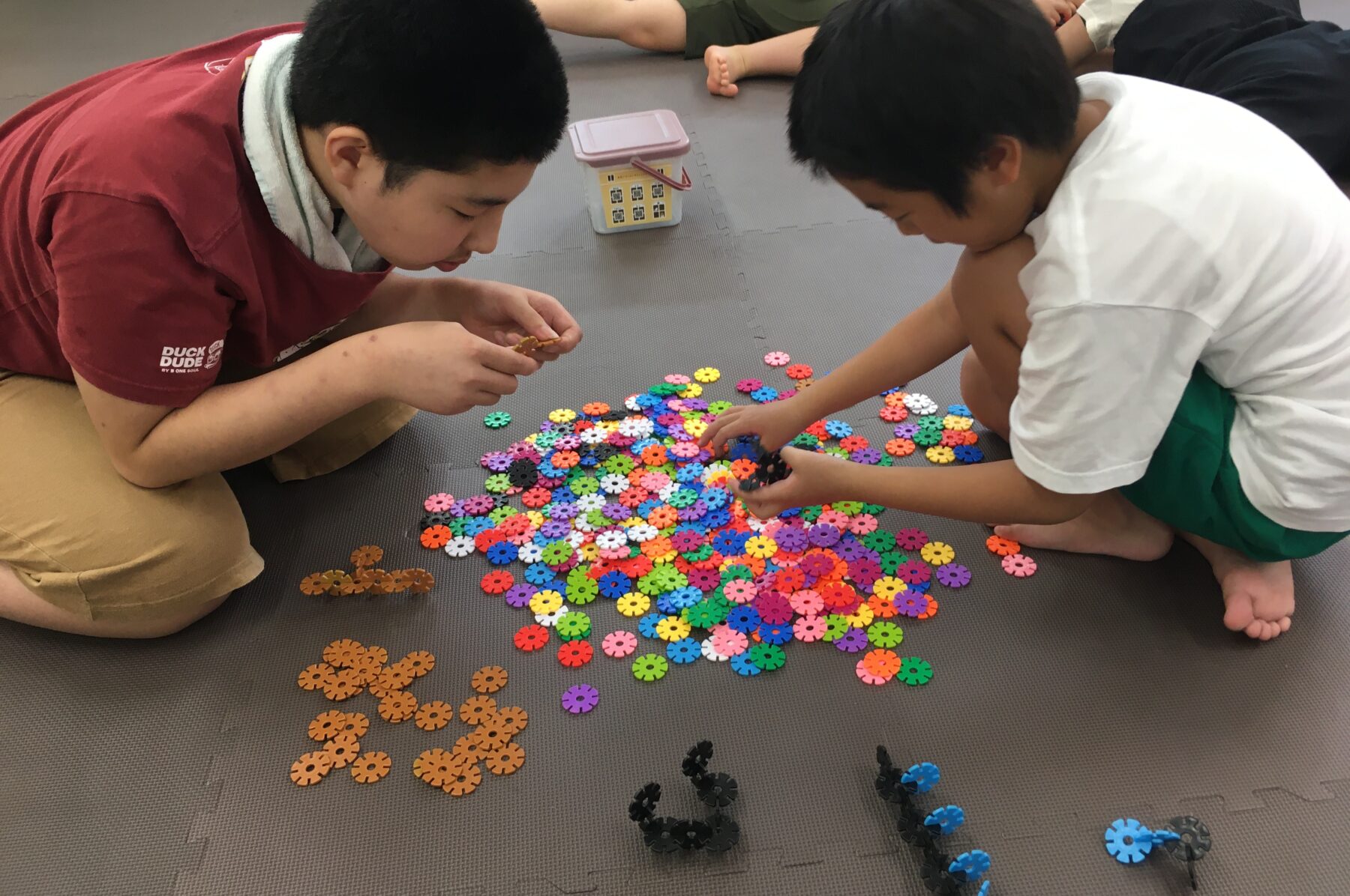 Two kids kneel on a grey mat, sorting a pile of colorful flower-shaped interlocking pieces for a construction toy.